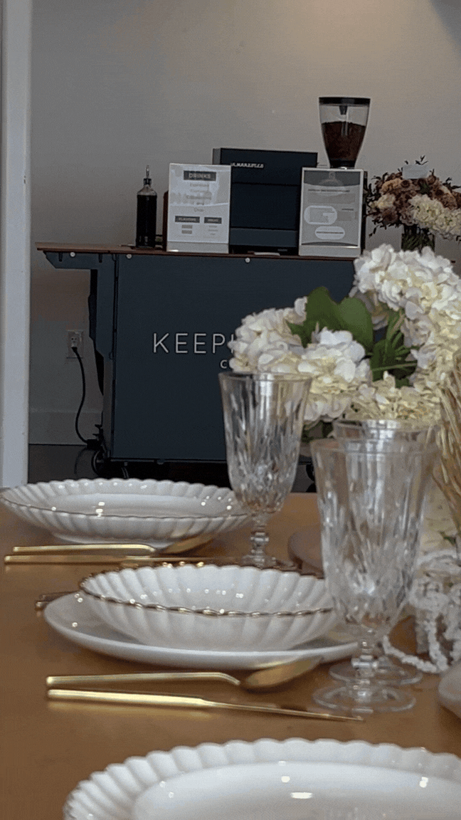 A dining table set with white scalloped plates, gold utensils, and crystal glasses, with a centerpiece of white flowers. In the background, there is a coffee station with a coffee machine, a glass jar filled with coffee beans, and a small bottle, all on a counter labeled 'KEEP...'.