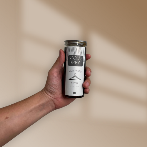 A hand holding a clear glass tumbler with a black lid, labeled 'Cold Brew Keep House Coffee.'