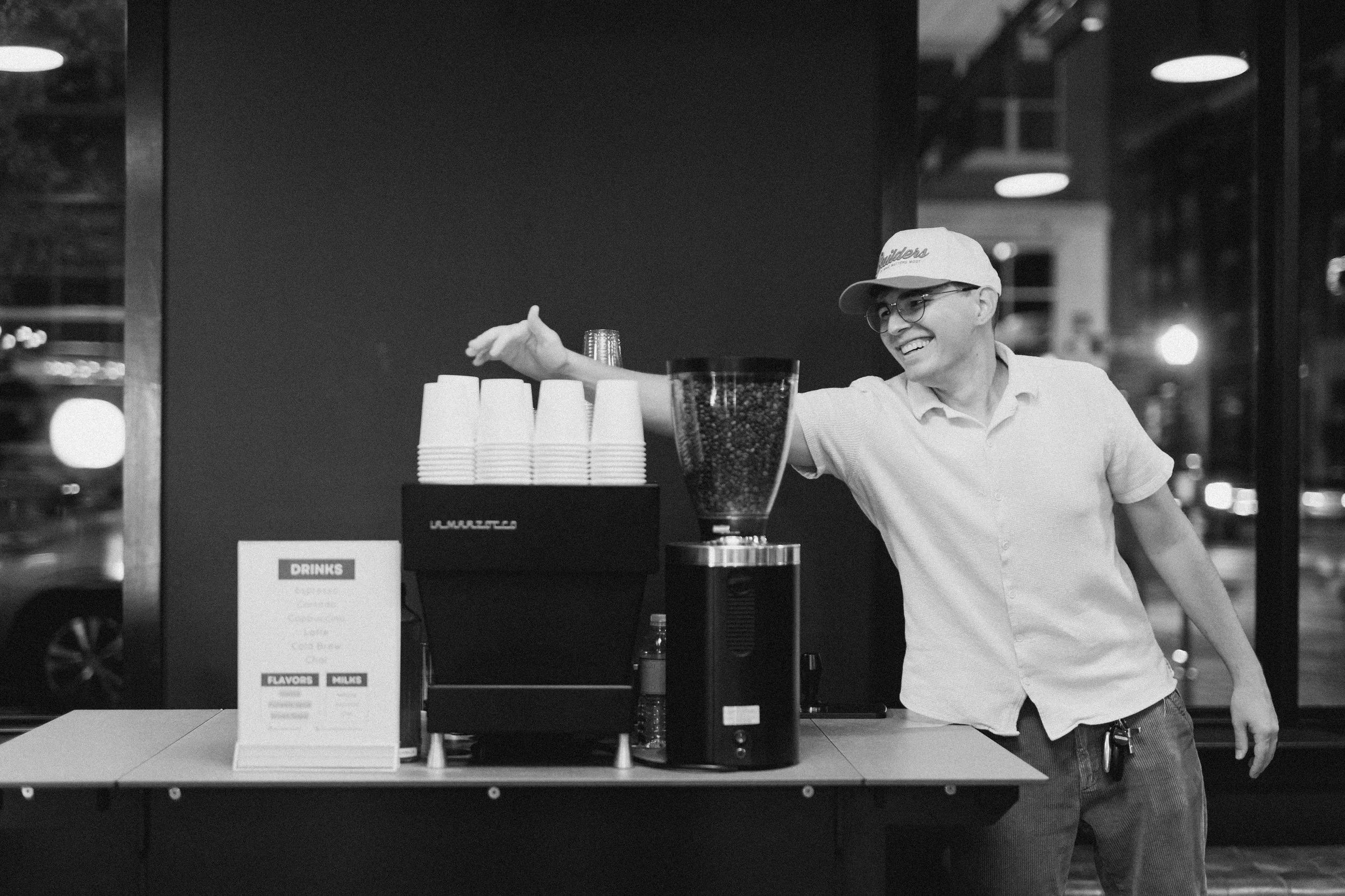 A smiling barista adjusting the espresso machine controls behind a wooden counter with a coffee grinder, a small plant, and a wristwatch.