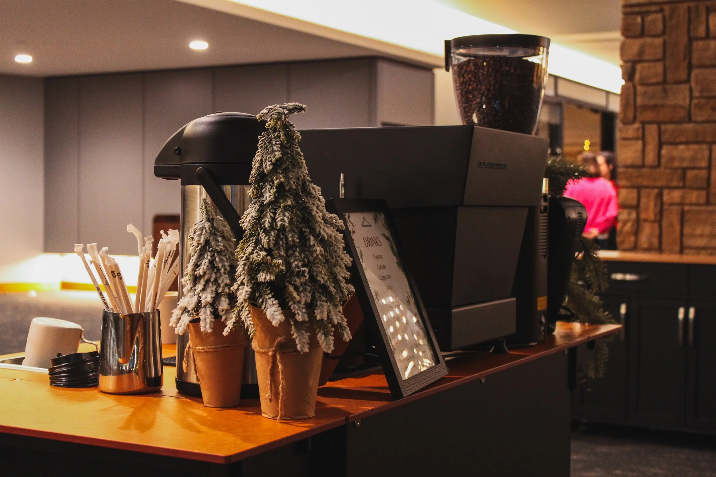 Coffee shop counter with Christmas decorations, including small snow-dusted pine trees in pots, a coffee machine, and a large jar of coffee beans.