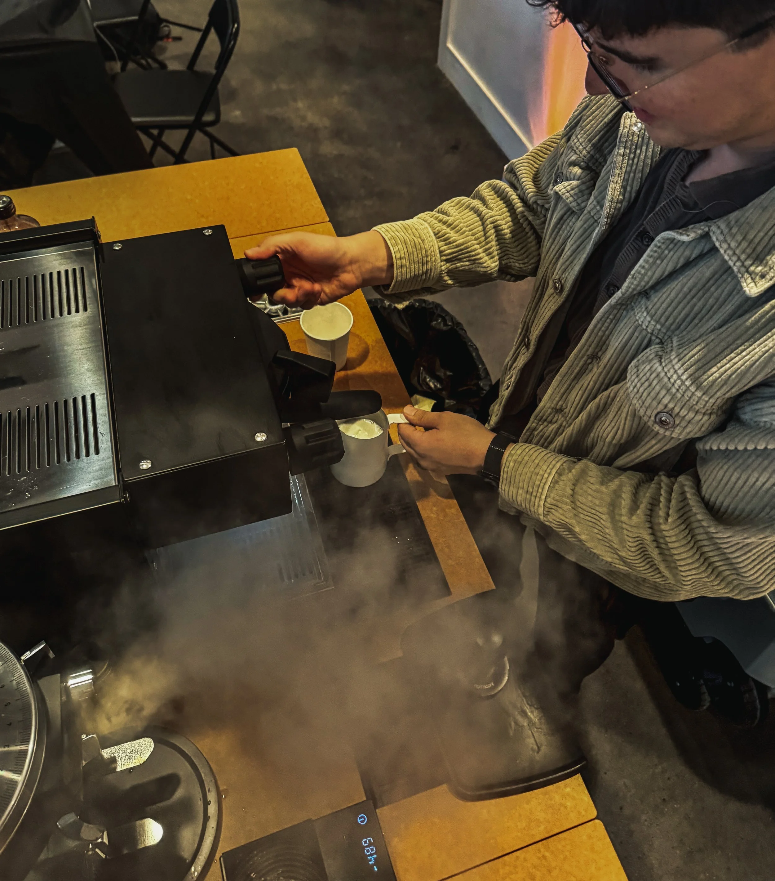A man is making coffee using a coffee machine at a wooden table with two cups, in a cozy indoor setting.