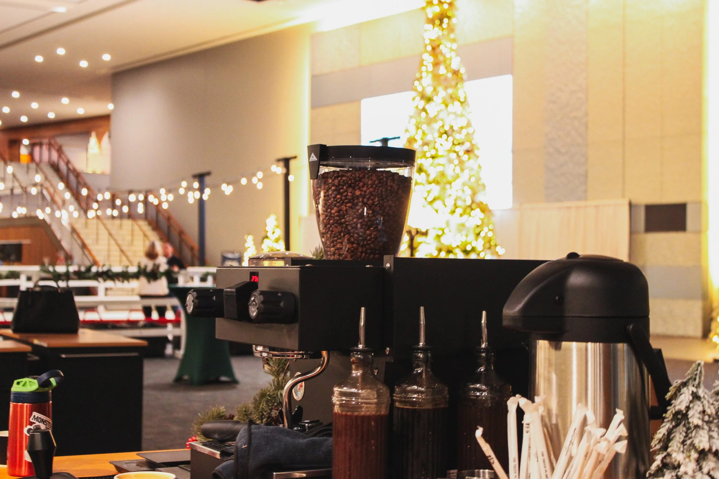 Coffee grinder with coffee beans, bottles of syrup, and a coffee carafe on a counter in a festive decorated room with Christmas trees and string lights.