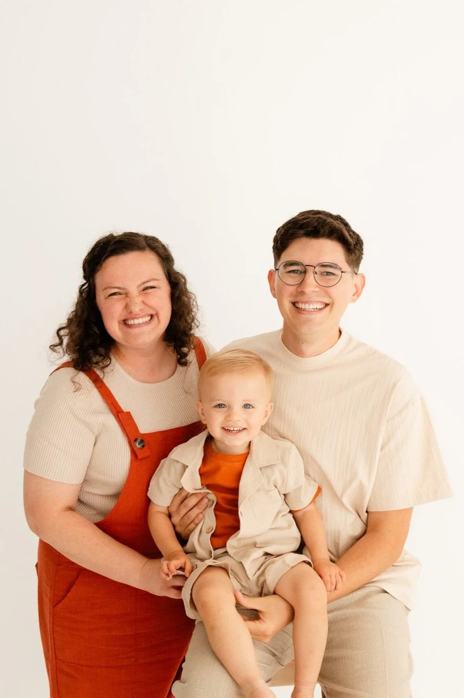 Smiling family of three with a woman, a man, and a young child, all wearing casual beige and orange clothing, against a plain white background.