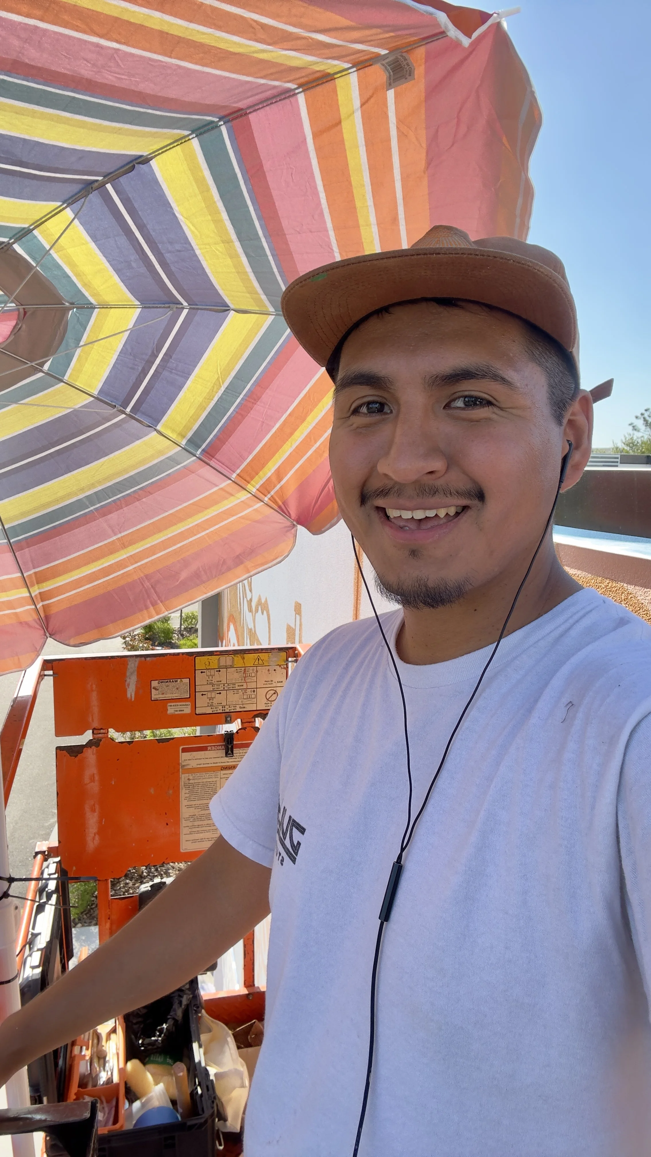 A smiling man wearing a tan cap, white t-shirt, and earphones, standing outdoors next to an orange equipment cart with a colorful striped umbrella overhead.
