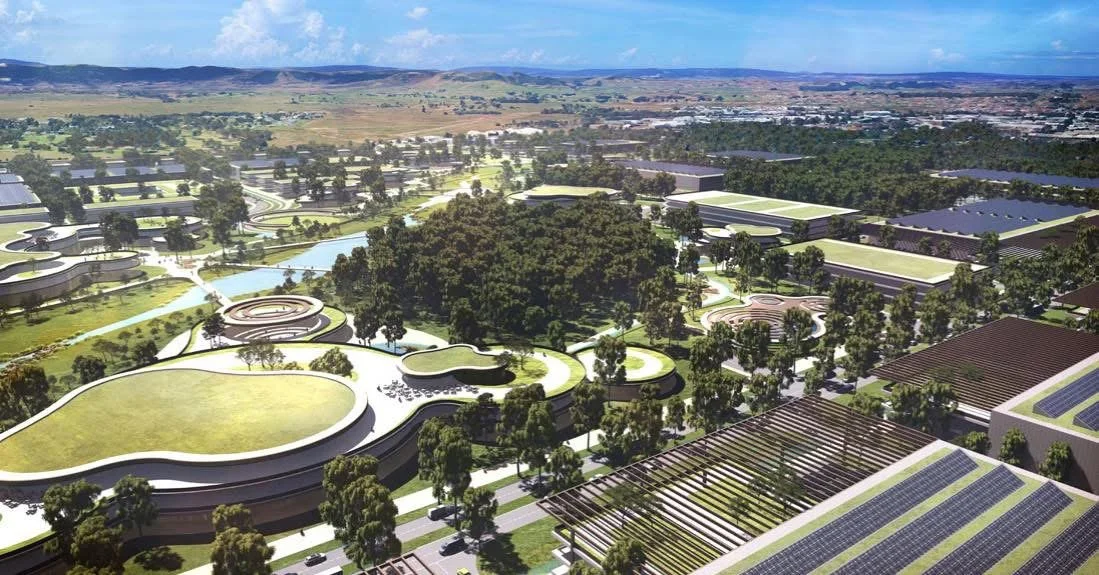 A large, modern urban park with multiple curved buildings, green roofs, and walking paths surrounded by numerous trees. In the background, there are more buildings, roads, and distant hills under a cloudy sky.