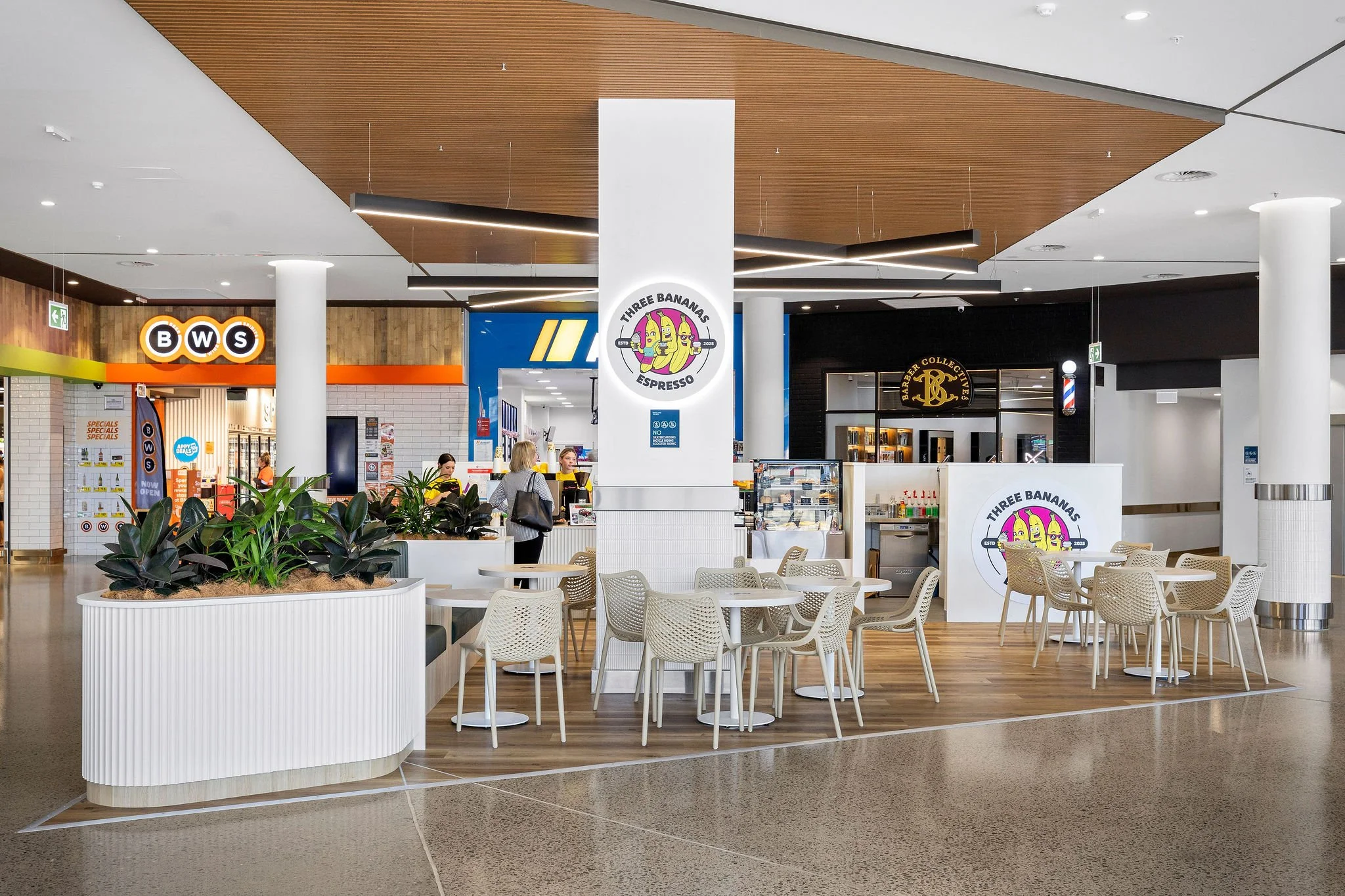 Interior of a modern shopping mall with a seating area. There are white tables and chairs, a large white planter with green plants, and various store signs including Three Bananas Espresso, BWS, and a barbershop. The ceiling features wood paneling and contemporary lighting.
