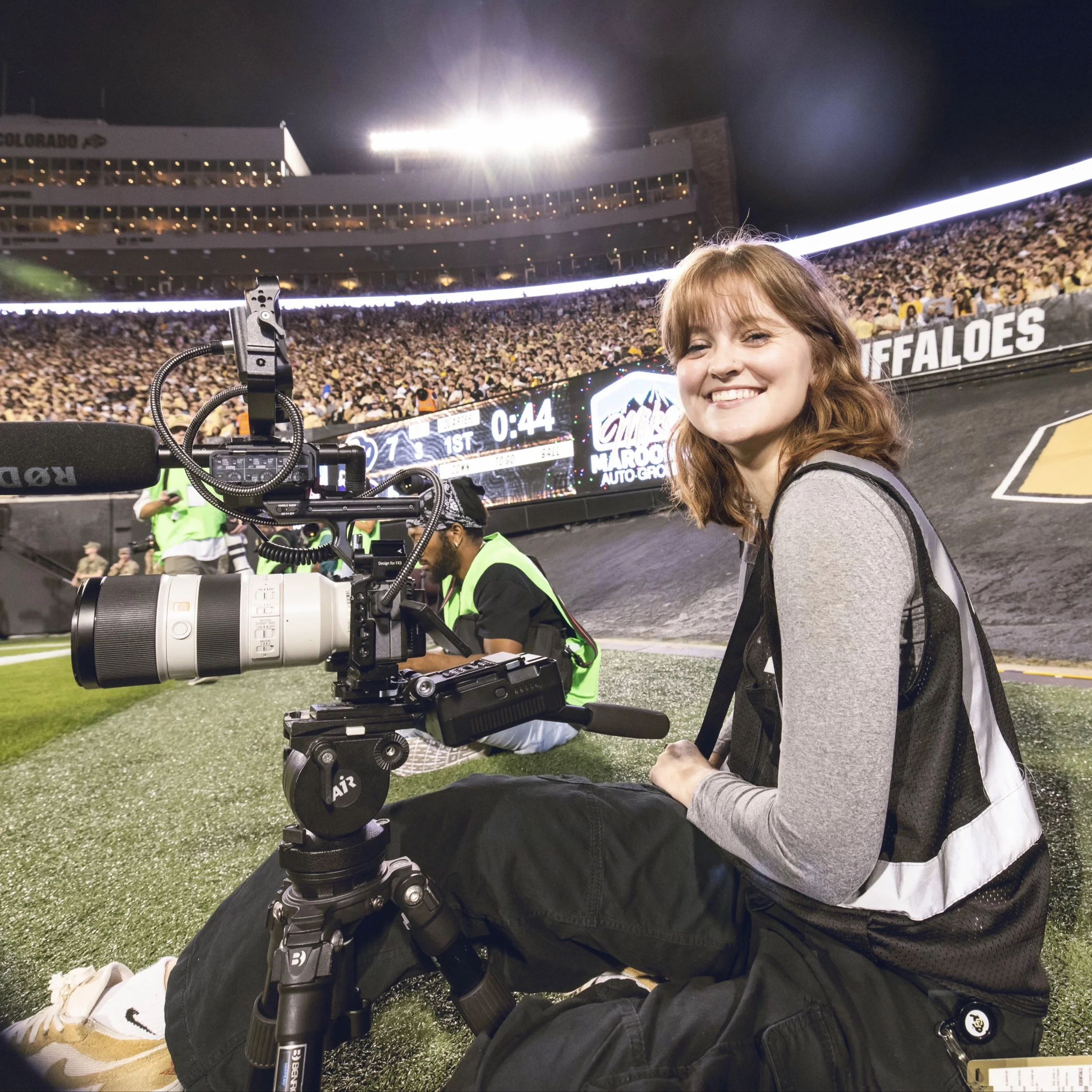 A young woman with red hair sitting on the ground at a sports stadium taking a break during a game, smiling at the camera with a large camera on a tripod in front of her. In the background, a man wearing a reflective vest is using a device, and the stadium is filled with spectators under bright lights with a scoreboard displaying 44 seconds remaining in the first quarter.