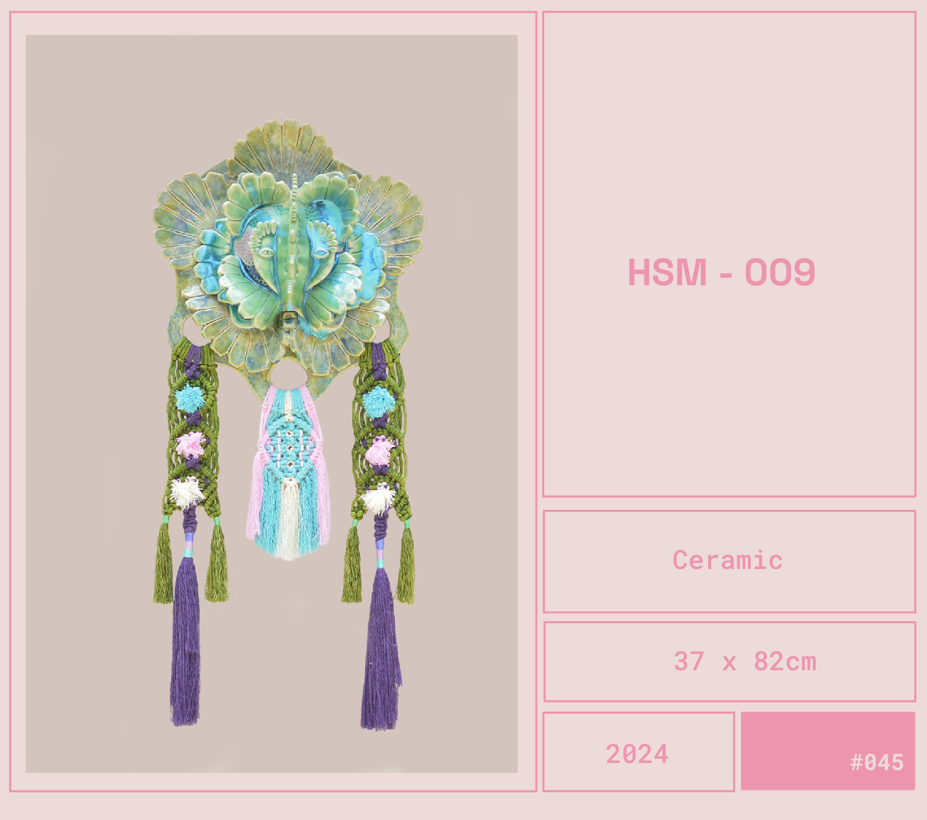 Una pieza de cerámica decorativa con un diseño inspirado en un rostro humano con elementos vegetales en tonos verdes y azules, con cordones de hilo y borlas en el centro y en los extremos.