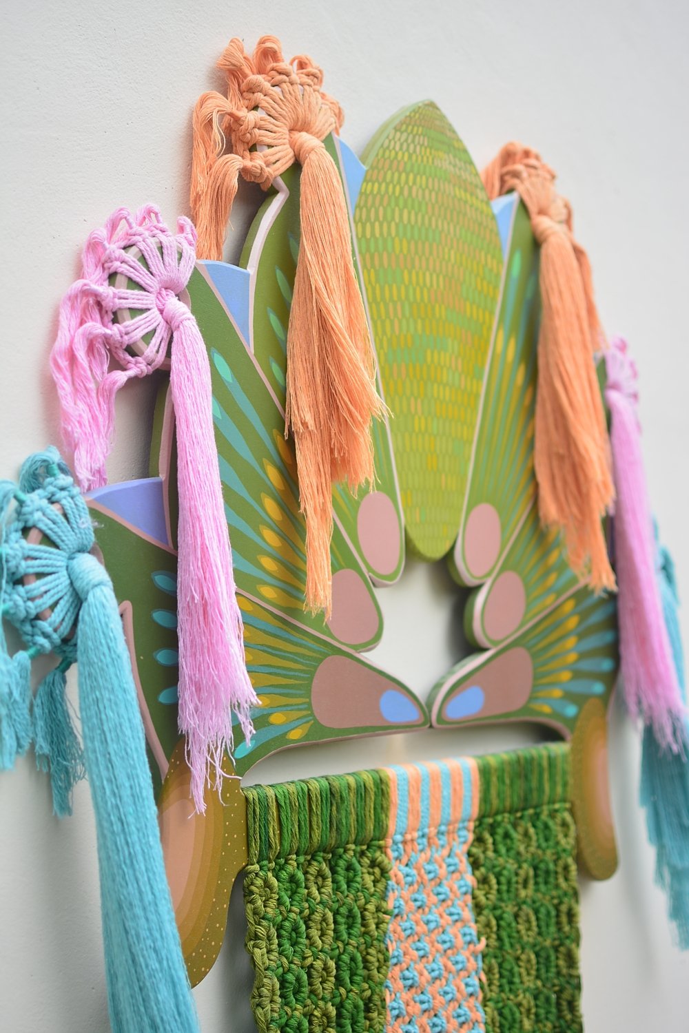 Decoración de pared con elementos coloridos, incluyendo hilos de colores en forma de flecos y tela tejida en tonos verdes, azules, rosas y naranjas, sobre un fondo de cartulina decorativa en forma de mariposa y formas geométricas.