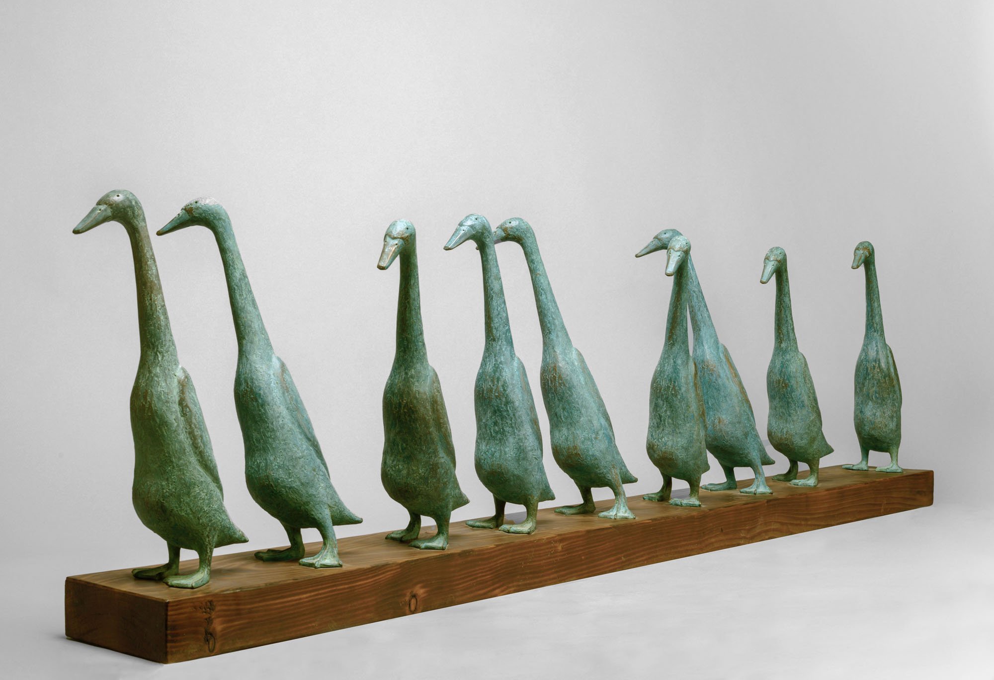 Seven metal duck sculptures with elongated necks and distorted bodies, mounted on a wooden base against a plain background.
