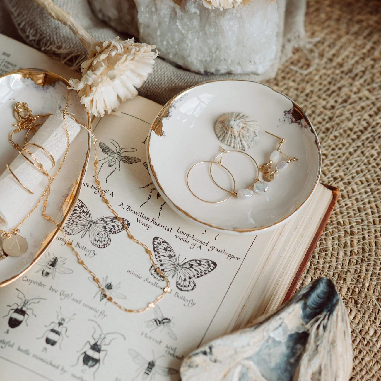 Jewelry displayed on a vintage book with butterfly and bee illustrations, surrounded by seashells, a DIY flower, and natural textured fabrics.