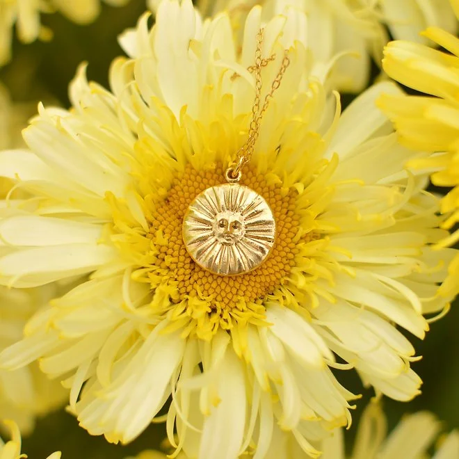 A gold sun pendant necklace with a smiling sun face hangs over a large yellow and white flower.