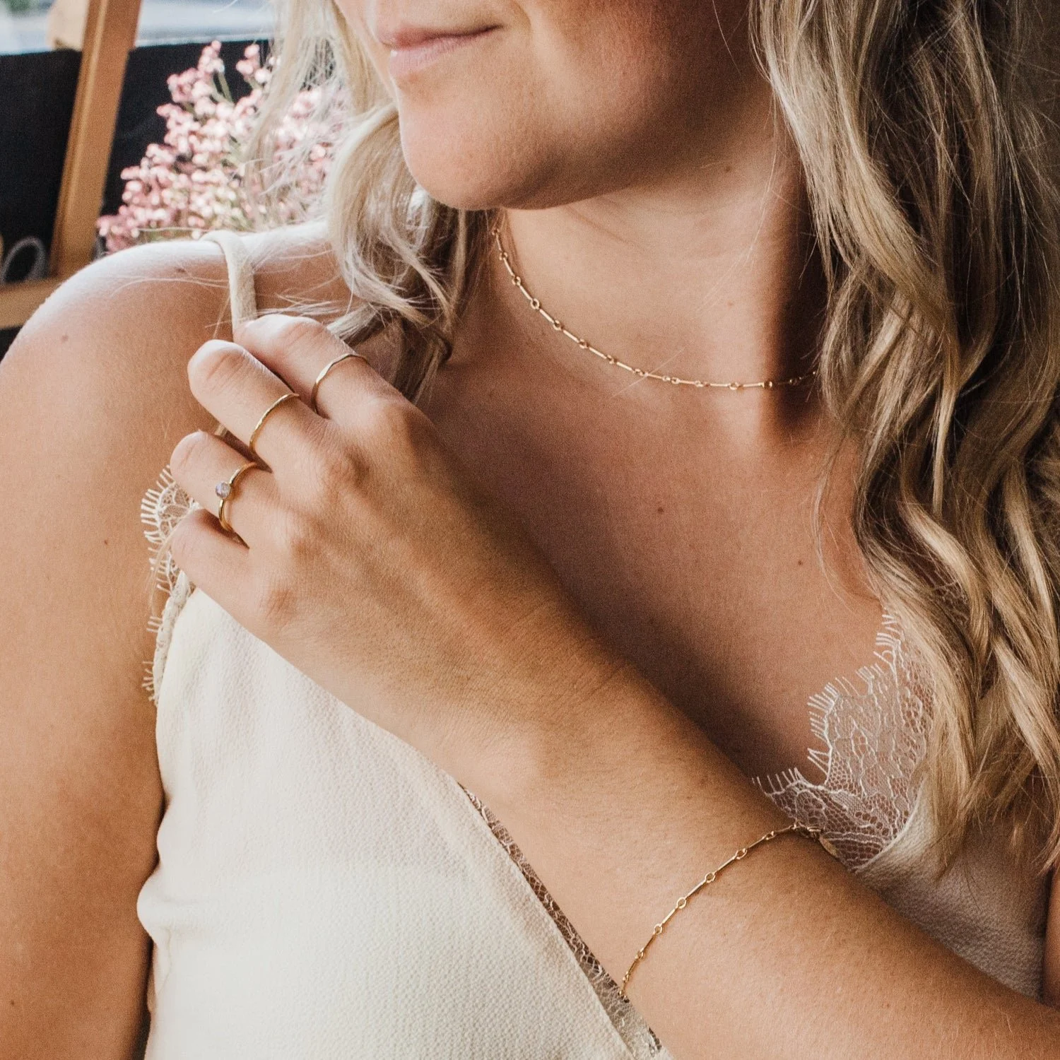 A woman wearing jewelry, including rings, a necklace, and a bracelet, touching her shoulder and wearing a cream-colored top with lace detail.