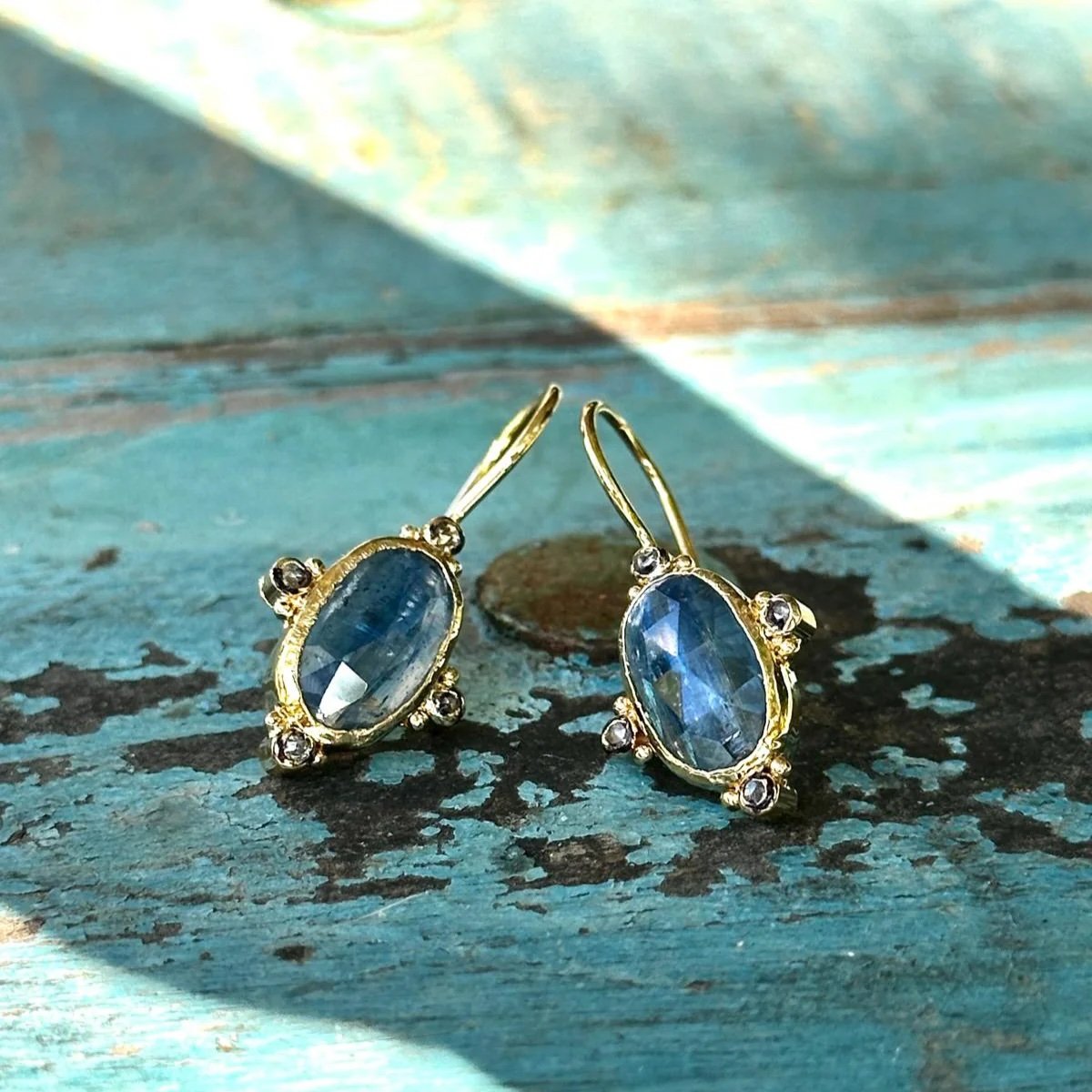 Gold earrings with oval blue gemstones and small surrounding clear stones, placed on a weathered blue wooden surface.