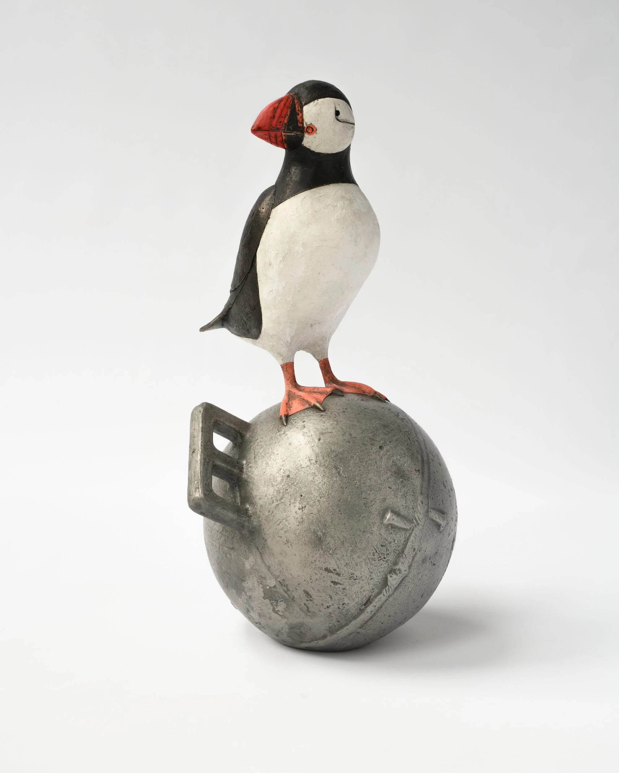 A sculpture of a puffin bird standing on a metallic bomb with a white background.