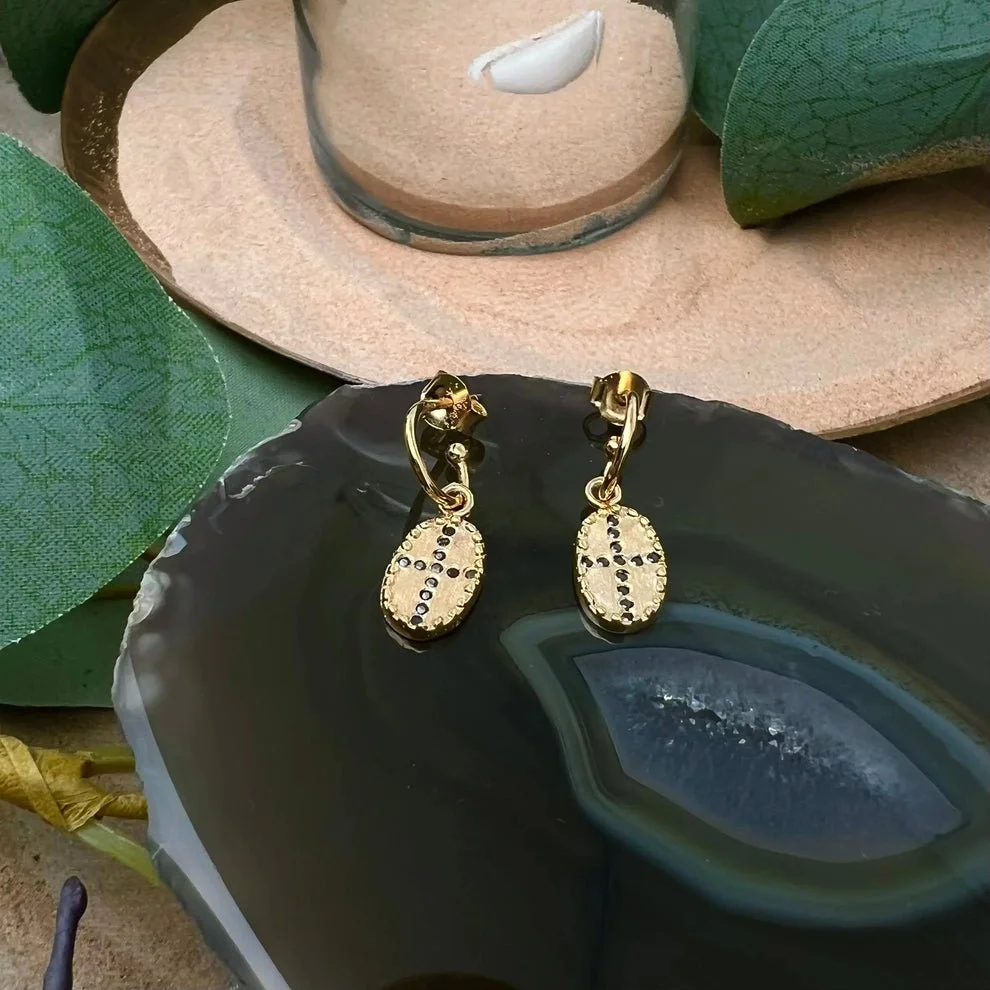 Gold hoop earrings with dangling oval pendants decorated with black stones, placed on a dark geode slice, with green leaves and a pinkish sand-colored hat in the background.
