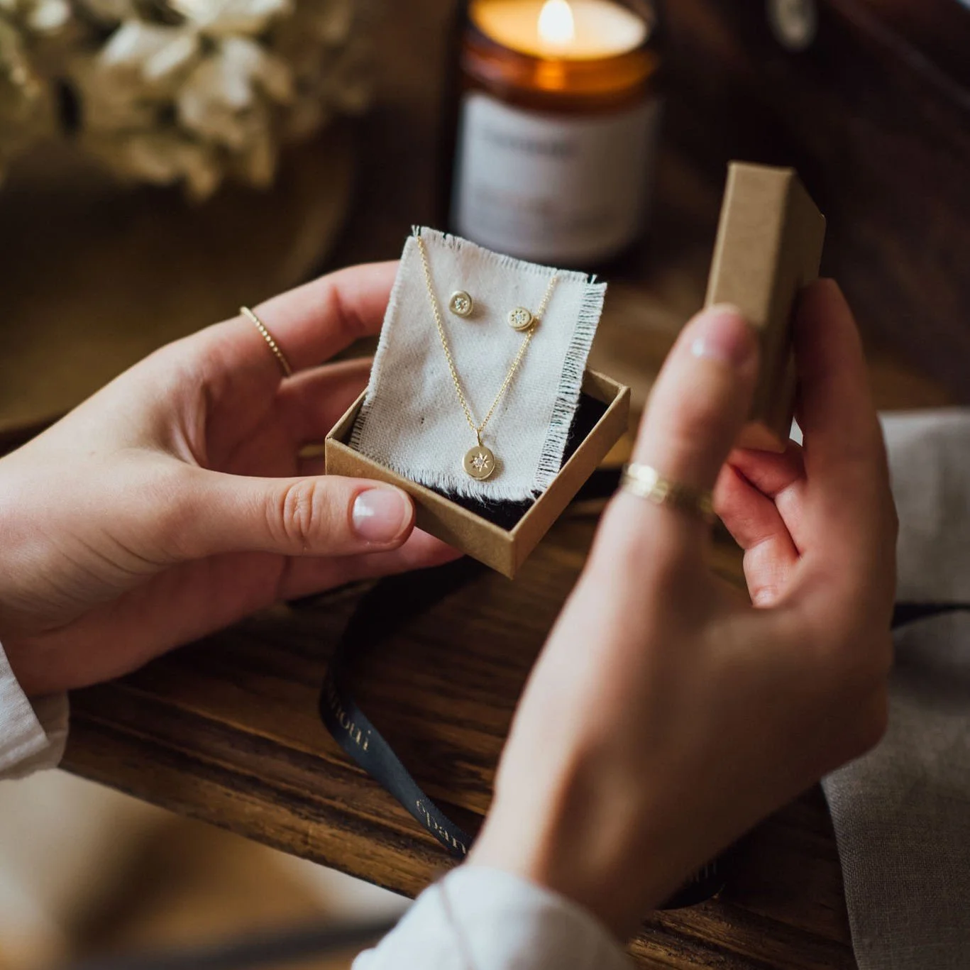 A person holding a small jewelry box containing a set of gold necklace and earrings with a snowflake charm, on a wooden table near a candle.
