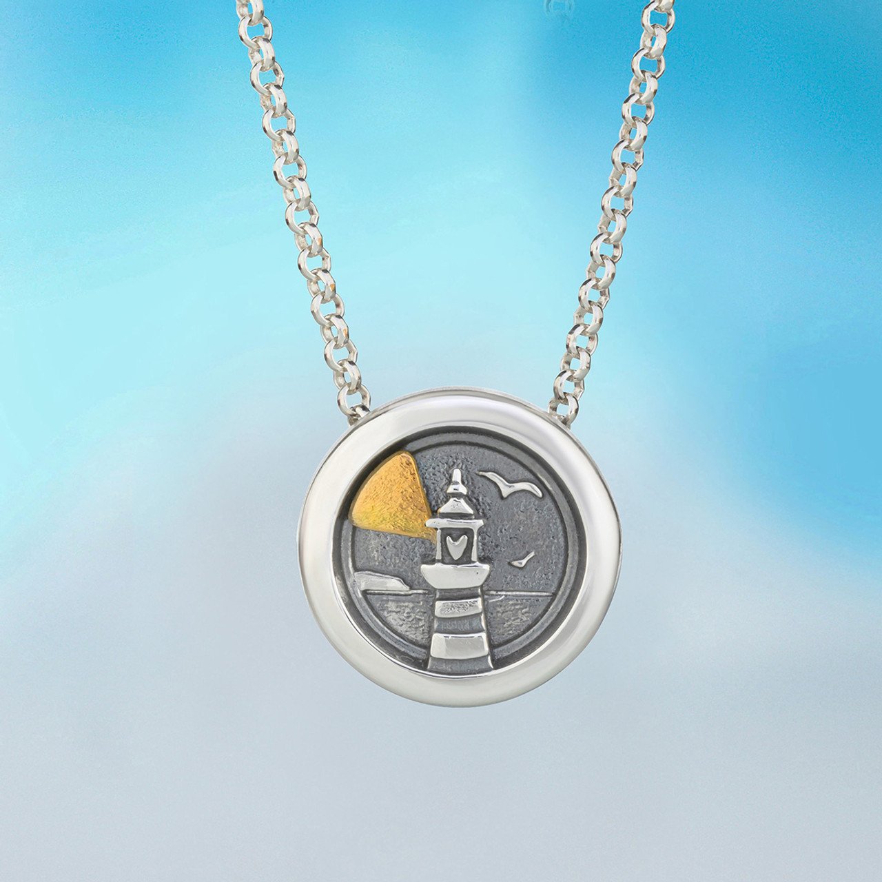 Silver necklace with a round pendant featuring a lighthouse scene, with part of the scene painted in gold.