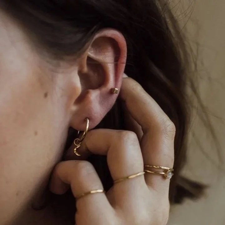 Close-up of a person's ear adorned with multiple gold earrings and rings on their fingers, with a hand touching the ear.