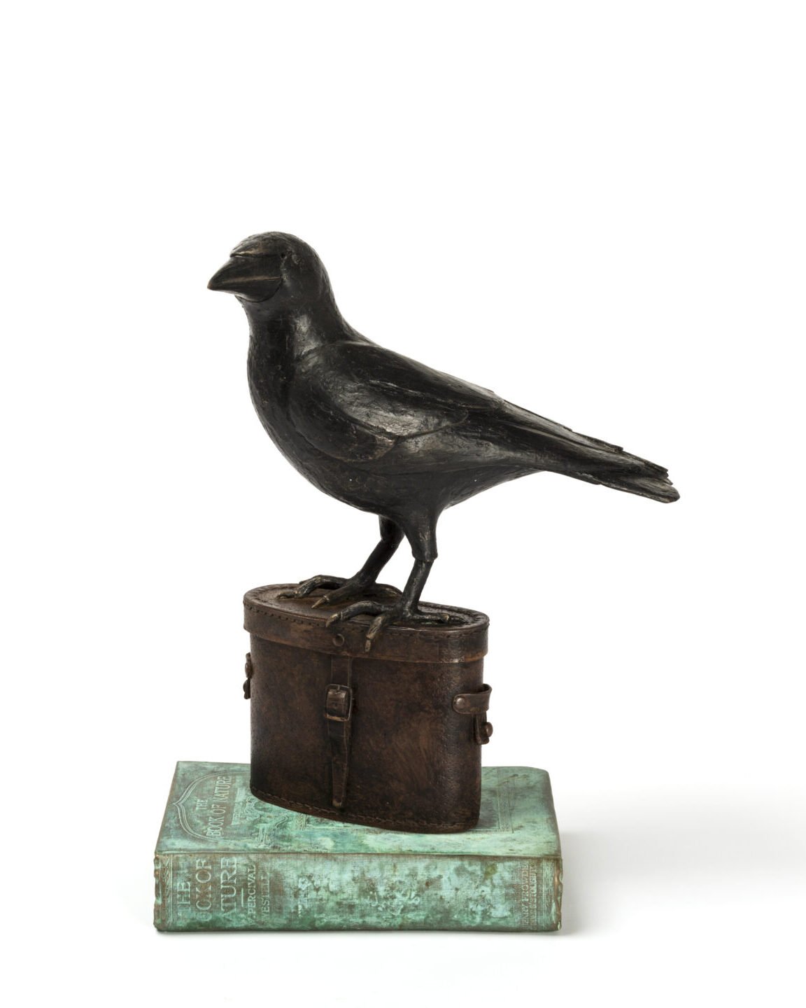 A black bird sculpture made of metal perched on a leather bag, placed on a worn green book.
