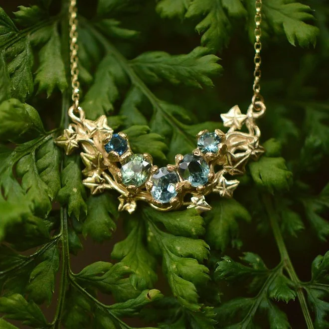 Gold necklace with blue gemstones and star-shaped accents hanging on a leafy green plant.