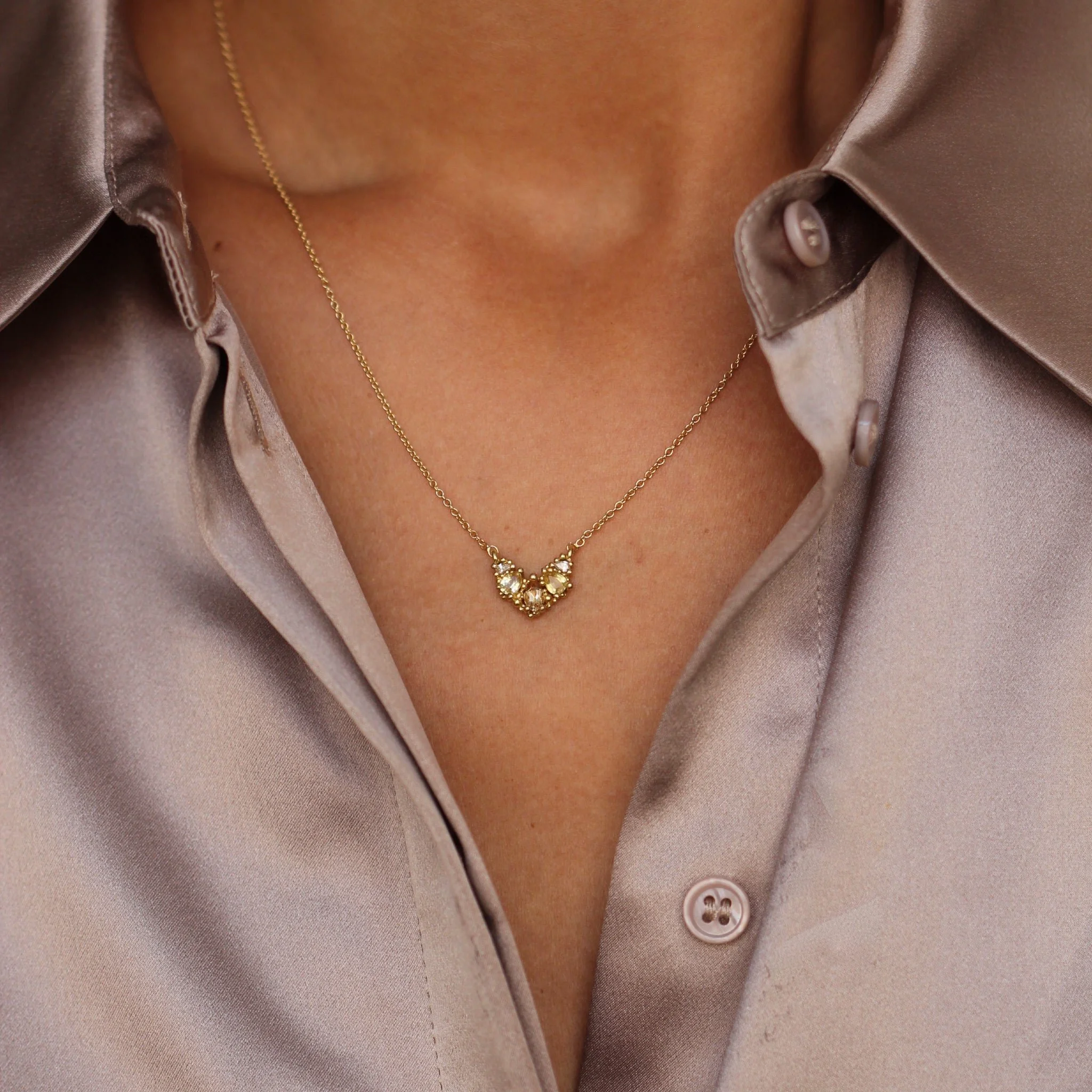 Close-up of a woman's neck wearing a gold necklace with a heart-shaped diamond pendant, and a satin beige button-up shirt.