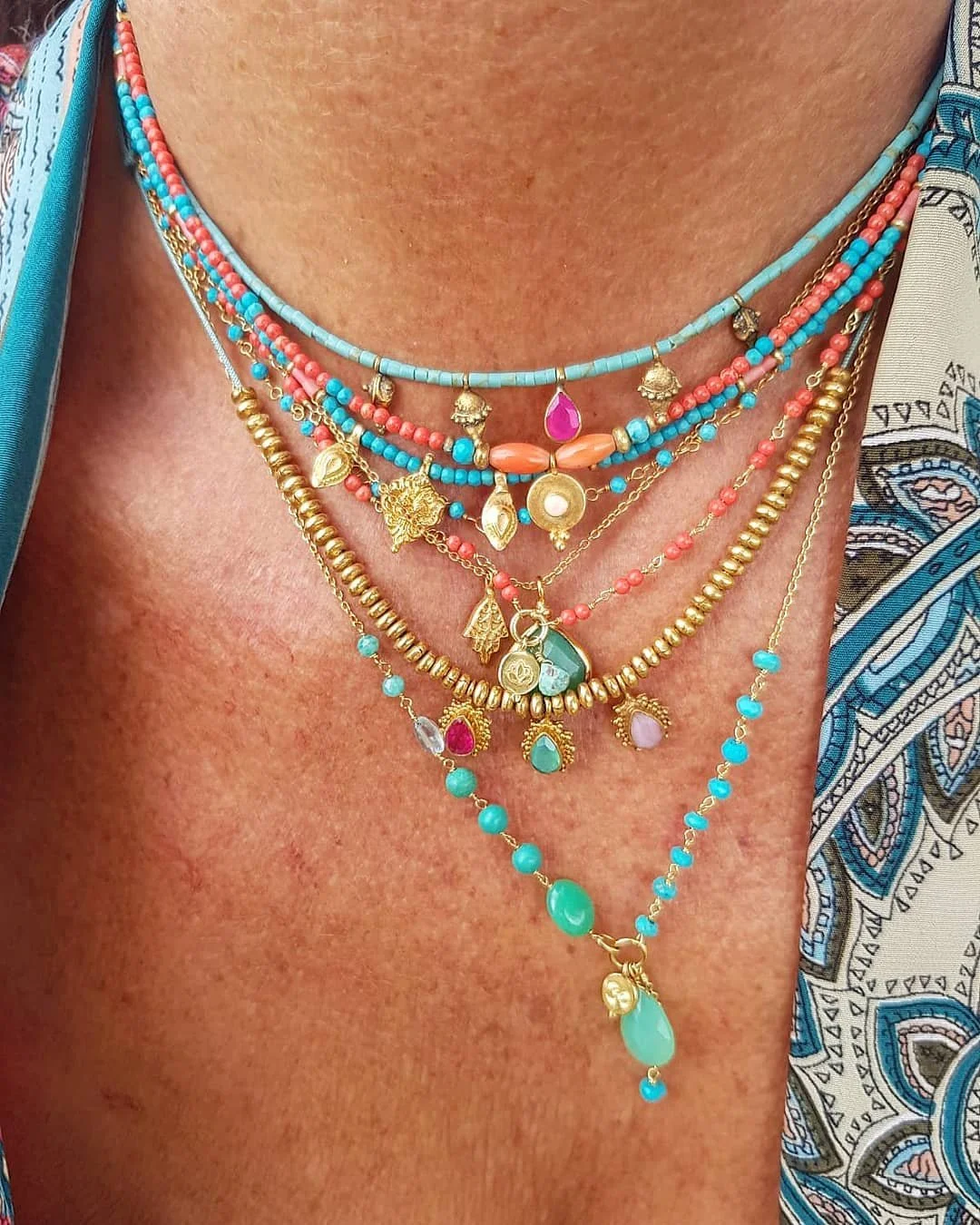 Multiple layered necklaces with colorful beads and gold charms on a person's neck, against a background of skin and patterned fabric.