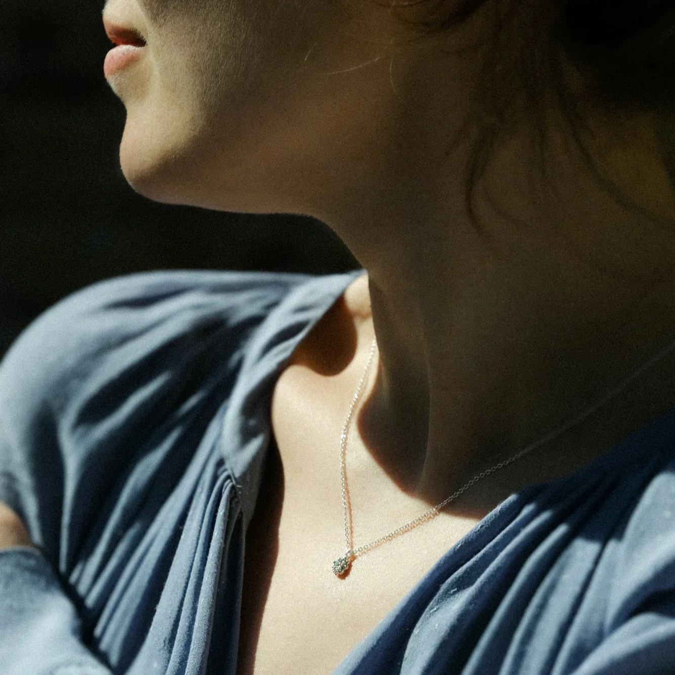 Close-up of a woman's neck and lower face, wearing a silver necklace with a small pendant, and a blue and white striped shirt or dress.
