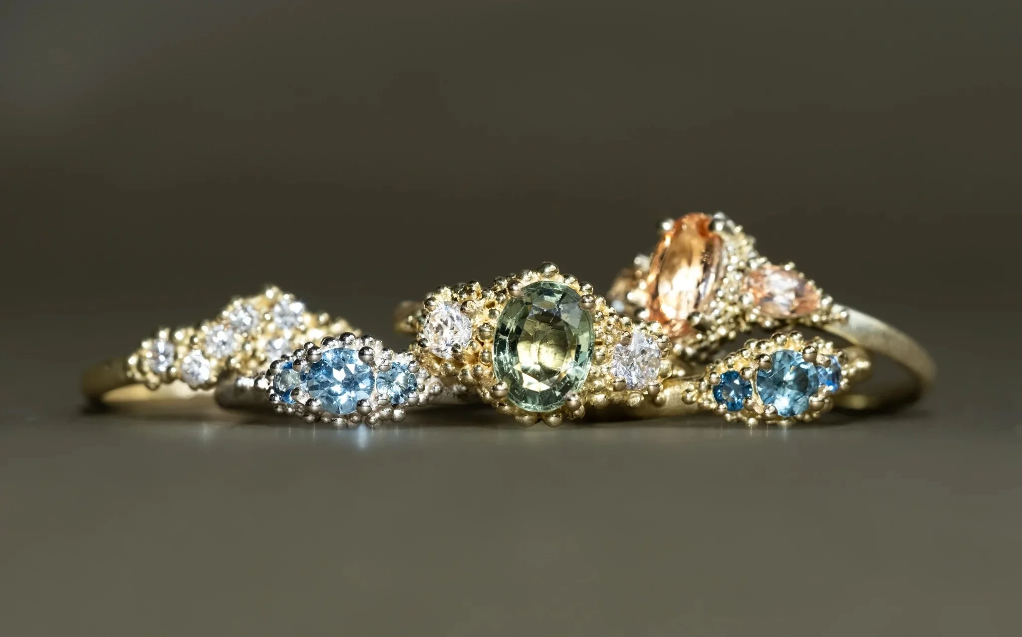 Collection of five gold rings with various gemstones, including blue, green, and clear stones, arranged on a dark surface.