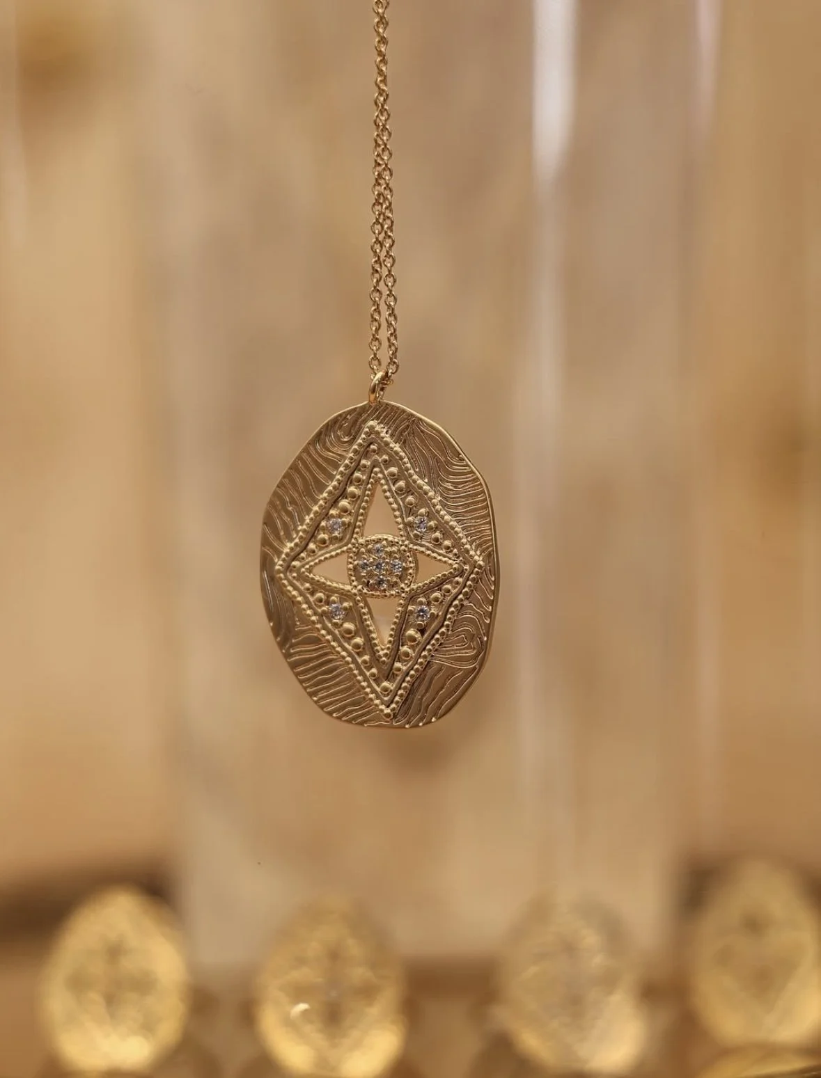 Gold pendant necklace with intricate geometric design and small gemstones, hanging in front of a blurred background of other jewelry pieces.