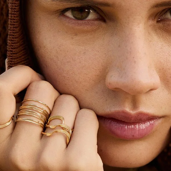 Close-up of a woman with brown eyes and light freckles, wearing multiple gold rings on her fingers, gently holding her face.