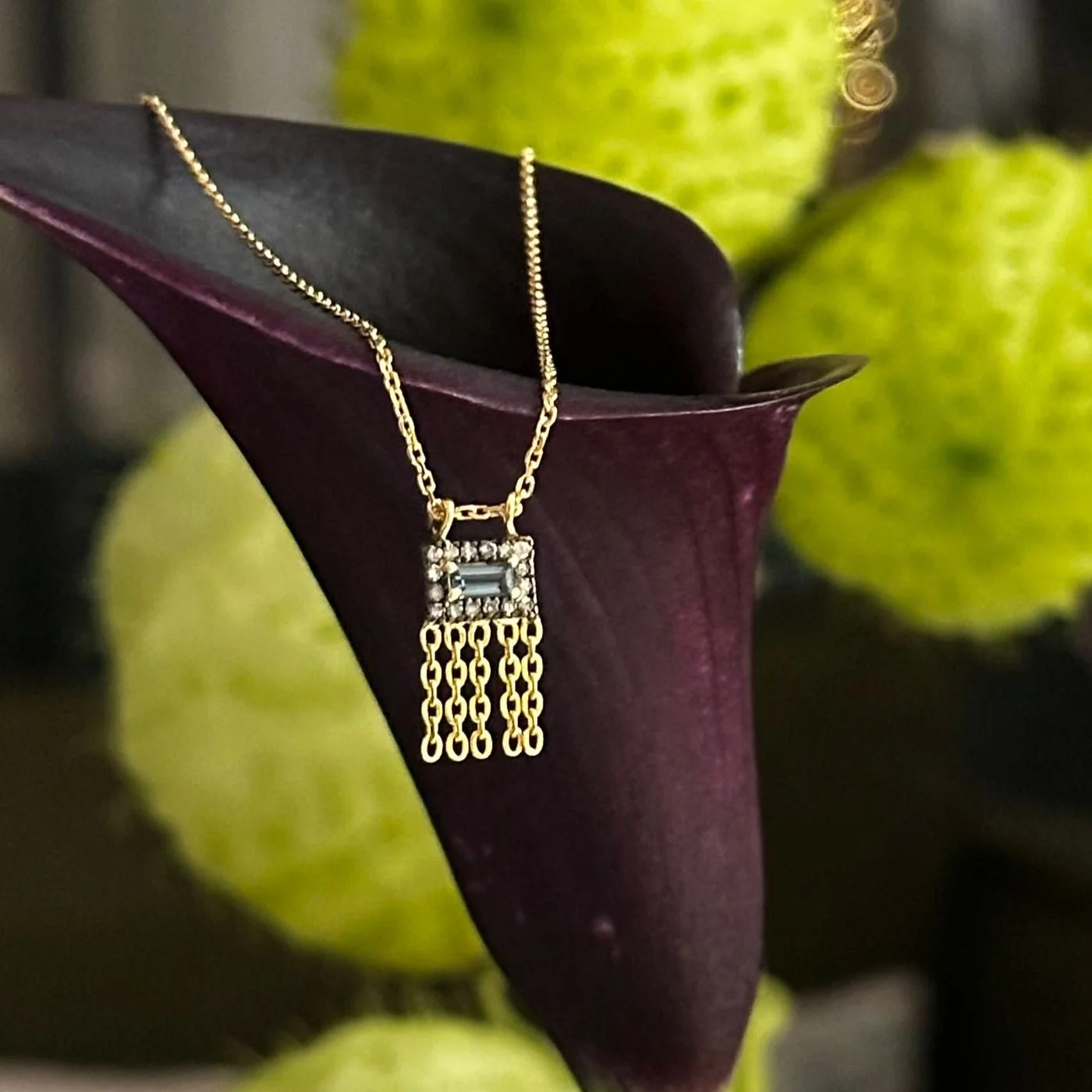 Gold necklace with a square pendant featuring a horizontal gemstone, hanging on a dark purple decorative vase with a green background.