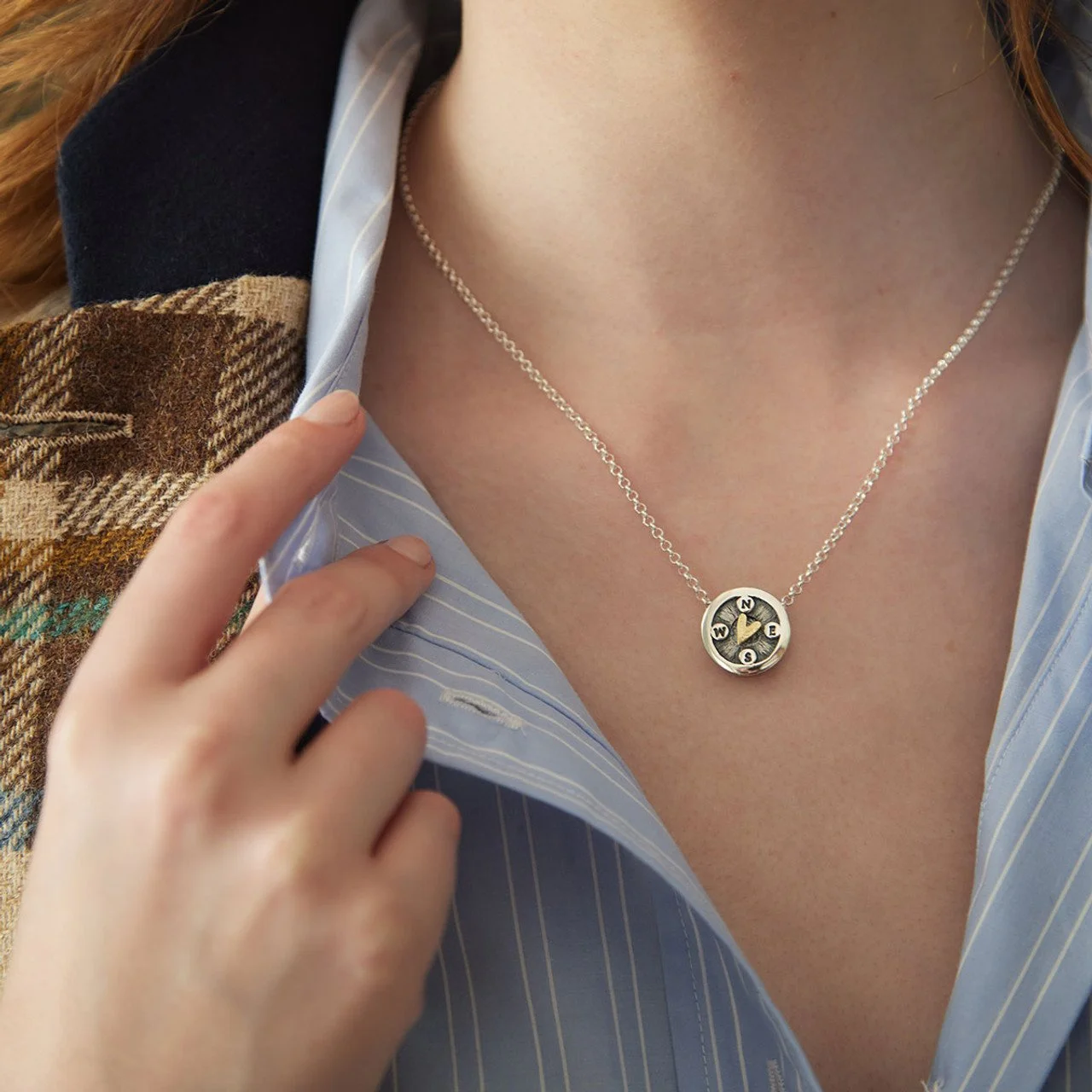 Person wearing a silver necklace with a compass design and a blue striped shirt, partially covered by a patterned outer shirt.