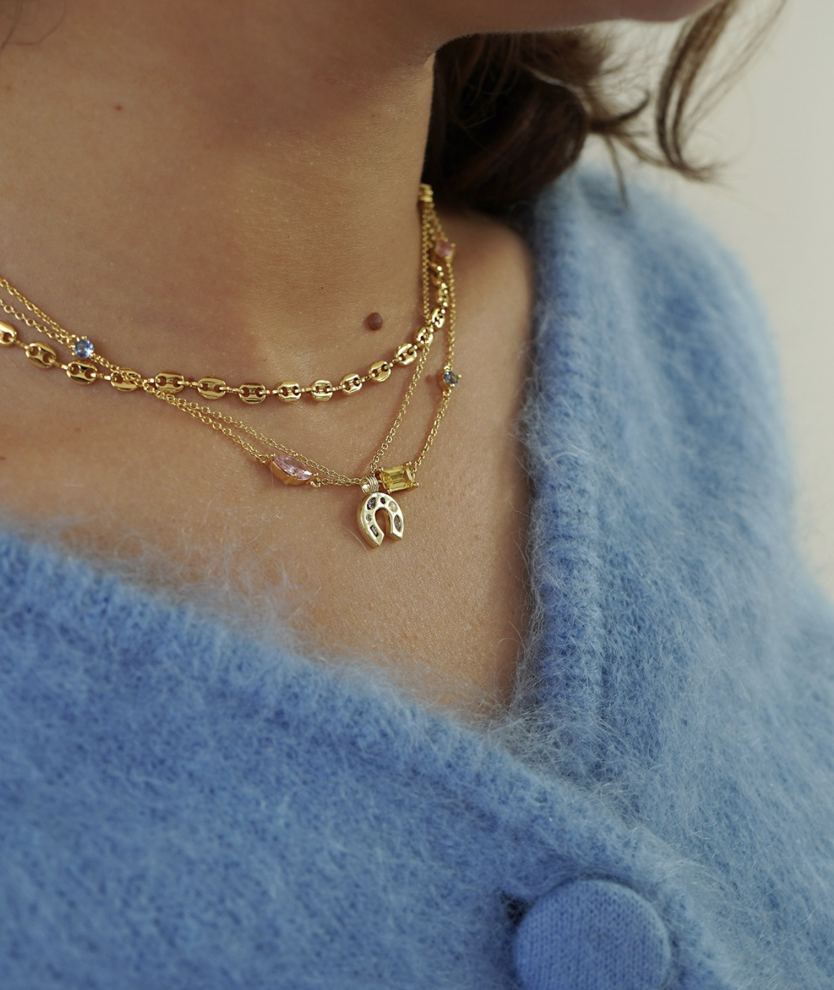 Close-up of a woman's neck and collarbone area wearing three layered gold necklaces with various pendants and charms, paired with a blue fuzzy sweater.