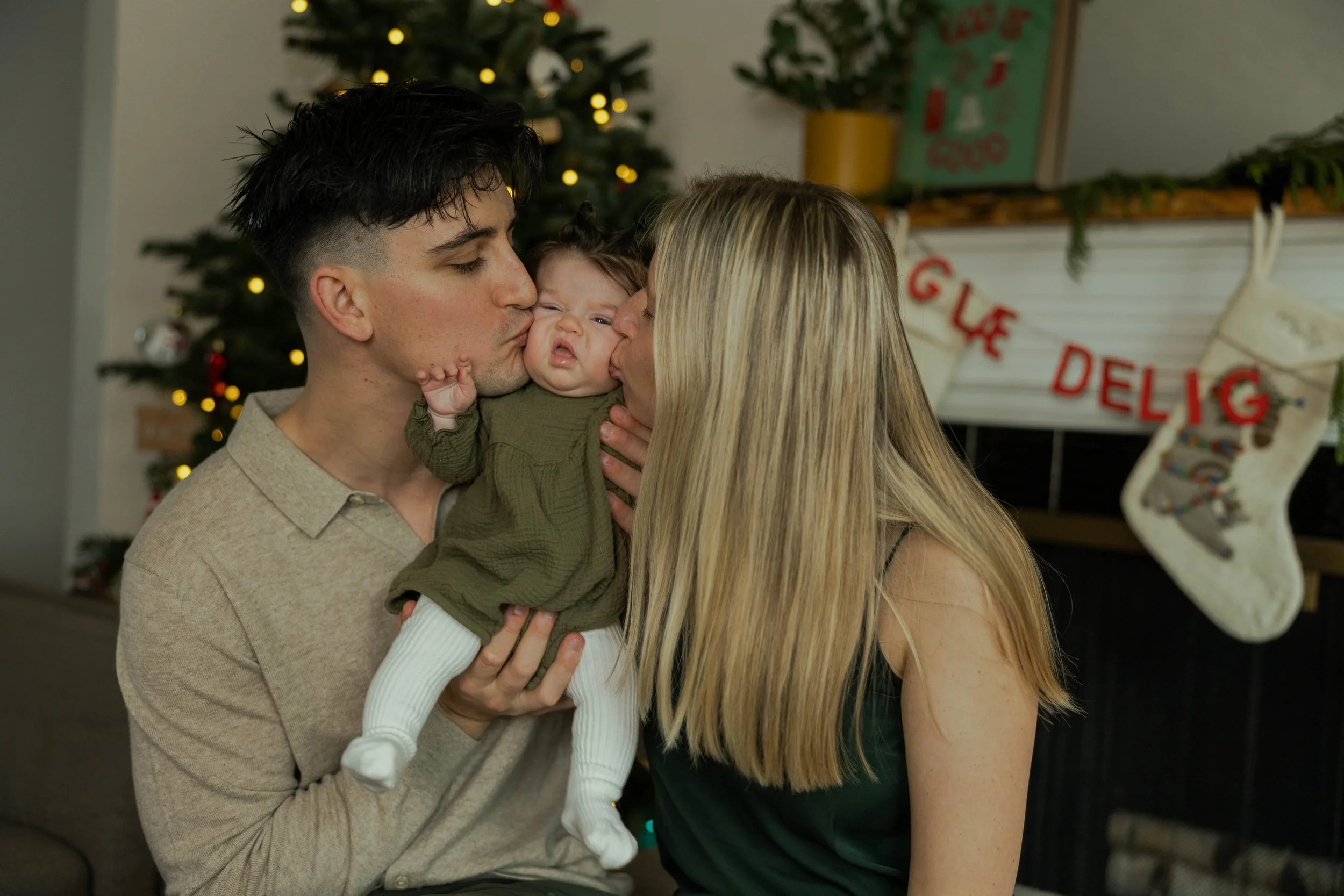 A family of three kissing during Christmas: a father, mother, and a baby daughter, with a decorated Christmas tree and stockings hanging in the background.
