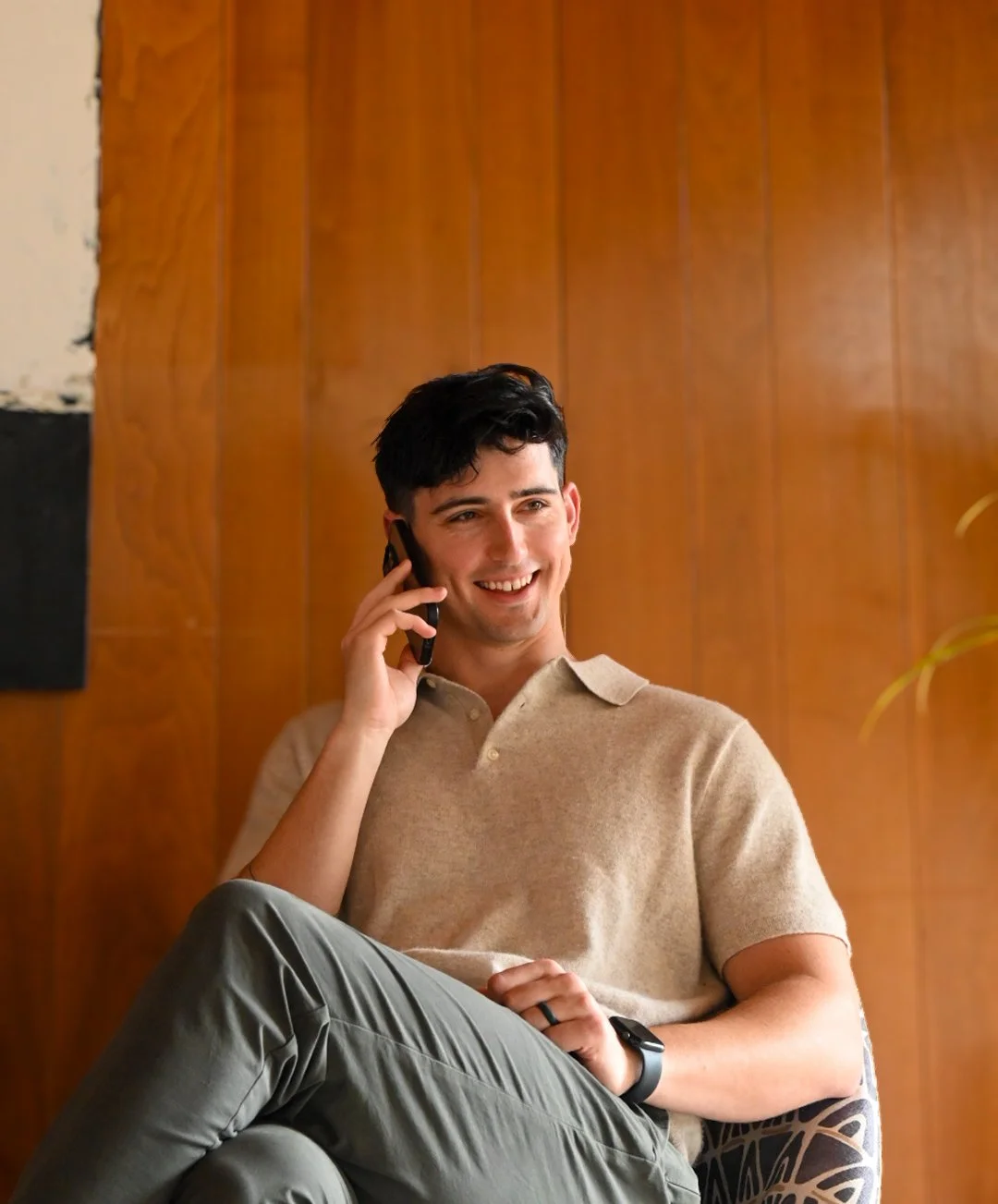 A young man with dark hair sitting on a chair, smiling and talking on his cellphone, wearing a beige polo shirt and gray pants, with a wooden wall in the background.