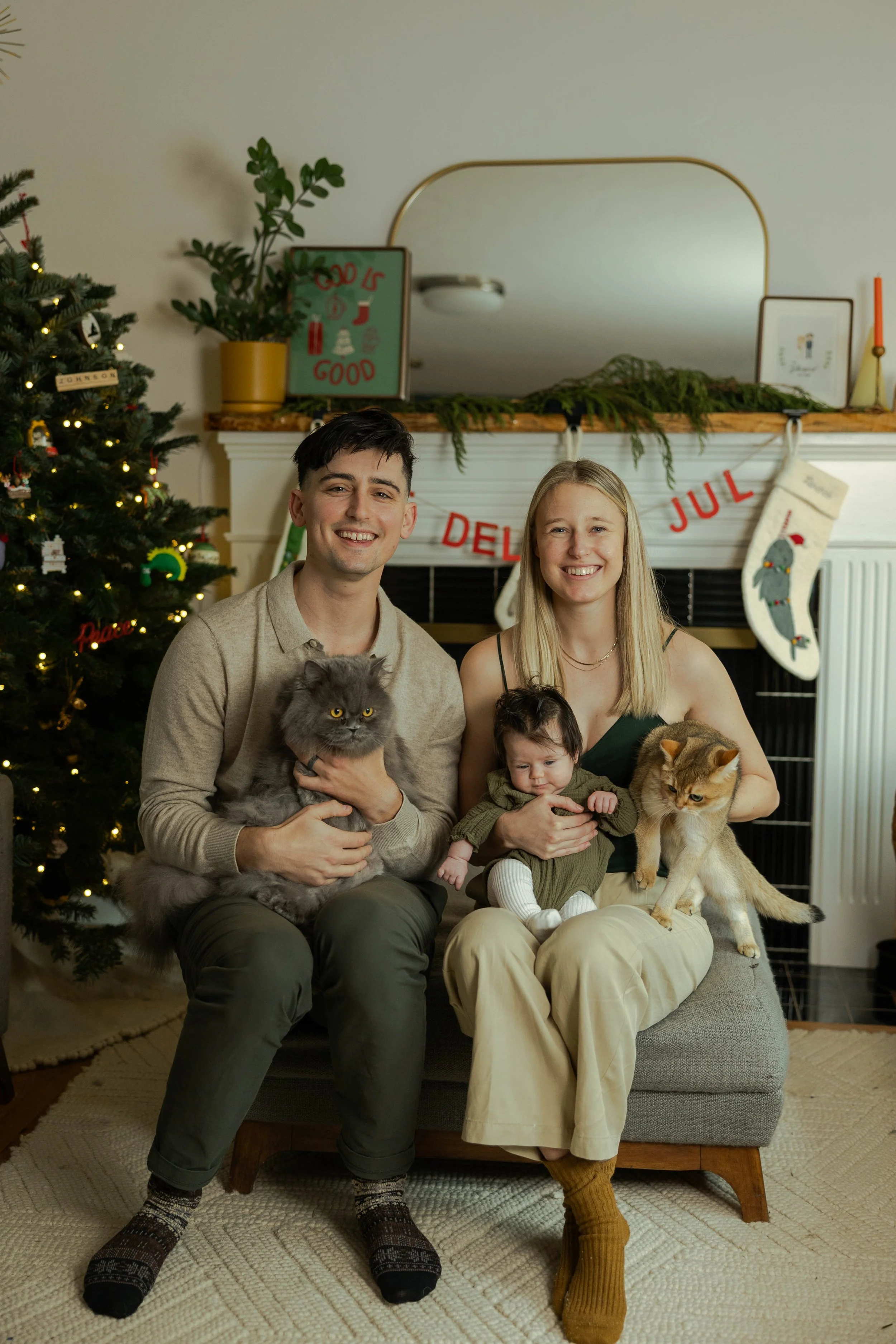 A family of four sitting on a gray bench in front of a decorated Christmas fireplace, holding their two cats and a baby. The Christmas tree is on the left with ornaments and lights, and holiday stockings hang on the mantel. A