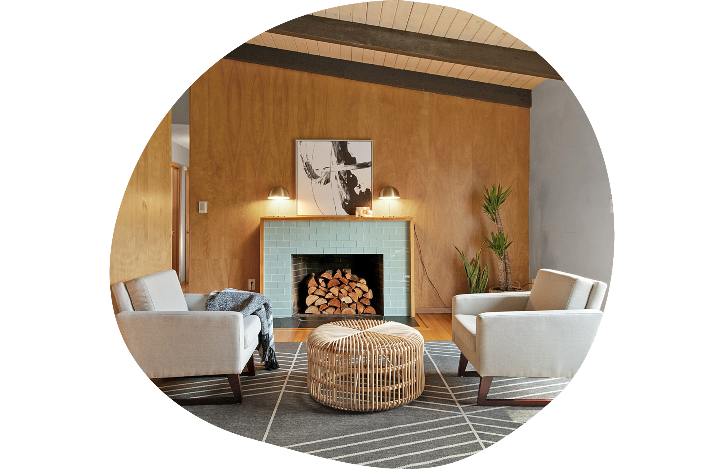 Living room with a wooden wall, a blue-tiled fireplace with firewood, two white armchairs, a round wicker coffee table, and potted plants, with a large abstract black-and-white artwork above the fireplace.