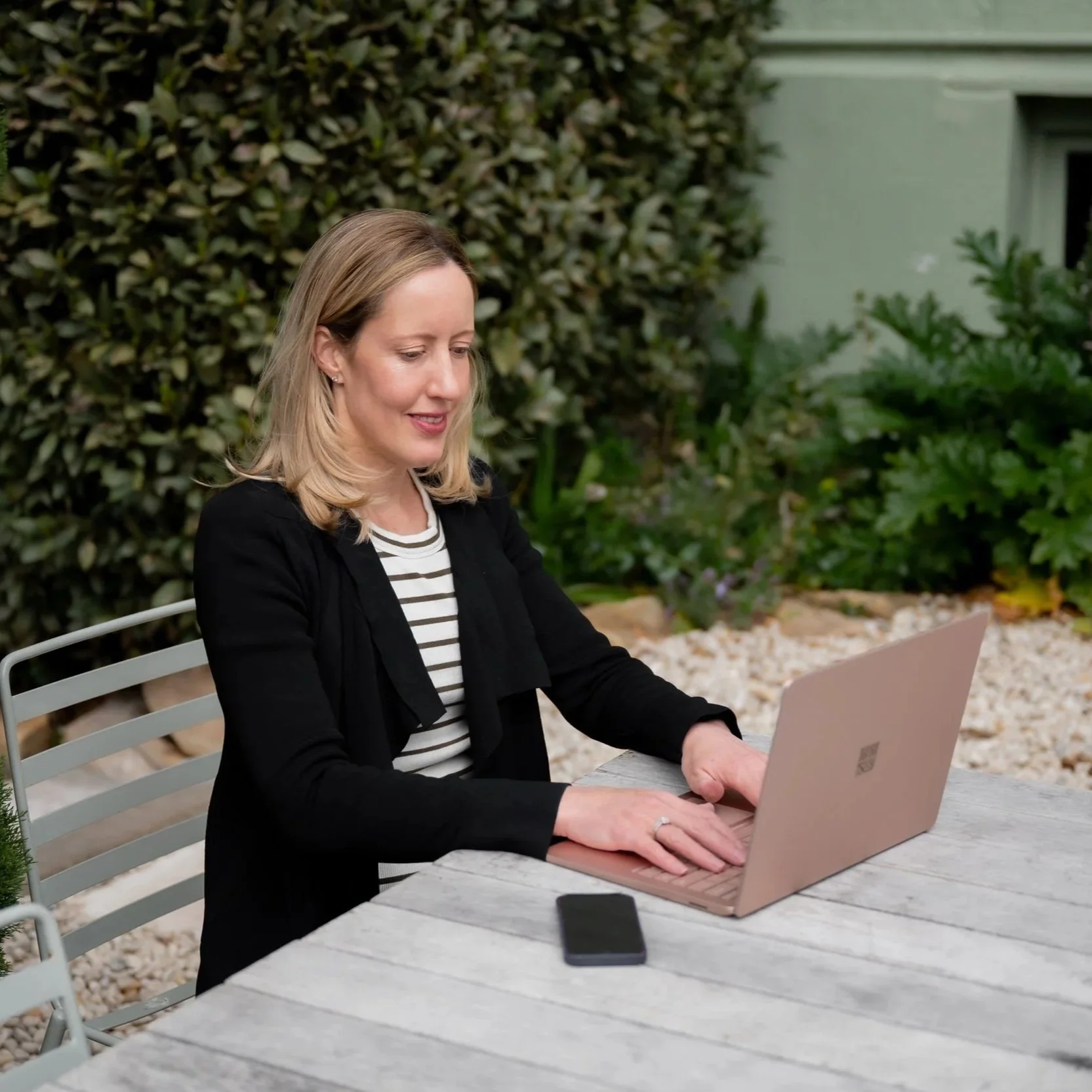A woman with shoulder-length blonde hair working on a pink laptop at an outdoor table surrounded by lush green plants and bushes.