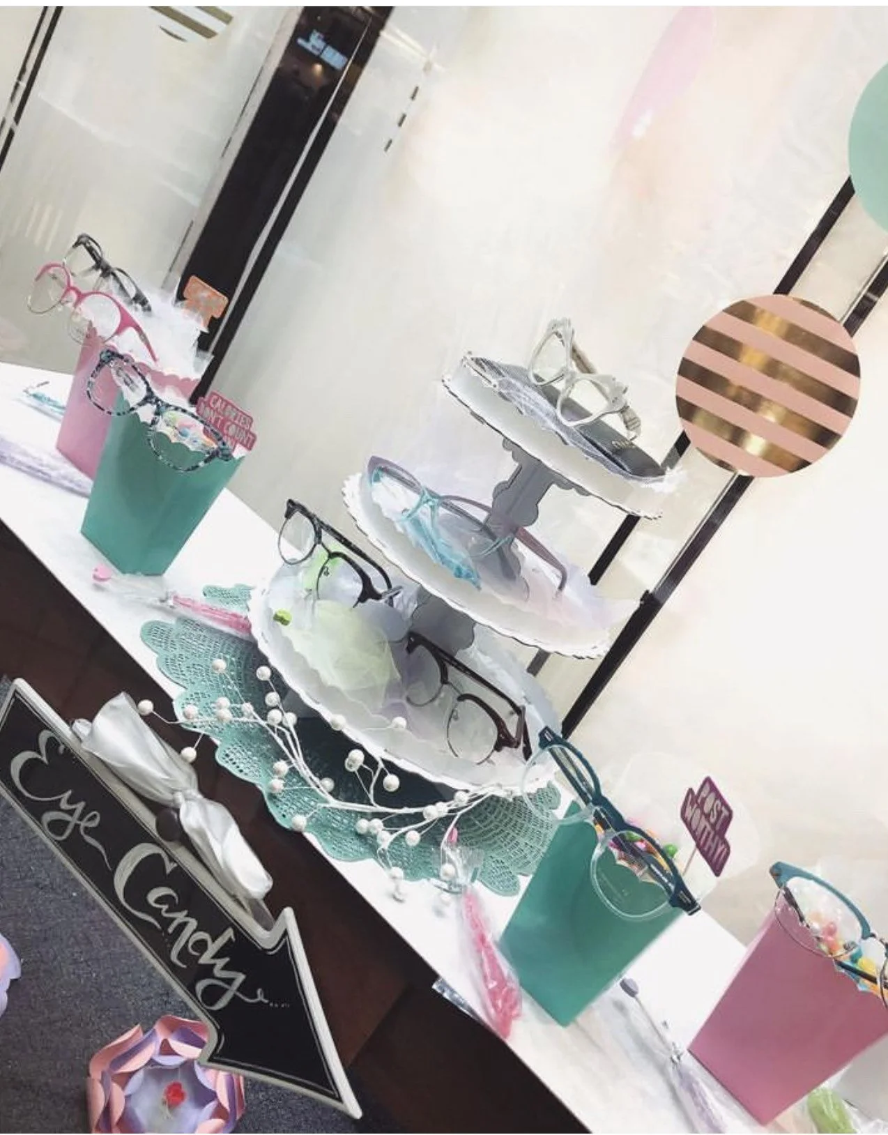 Display of colorful eyeglasses on tiered white trays and in pastel-colored buckets, decorated with a sign that says 'Eye Candy', and floral and decorative accents on a table at a retail store.