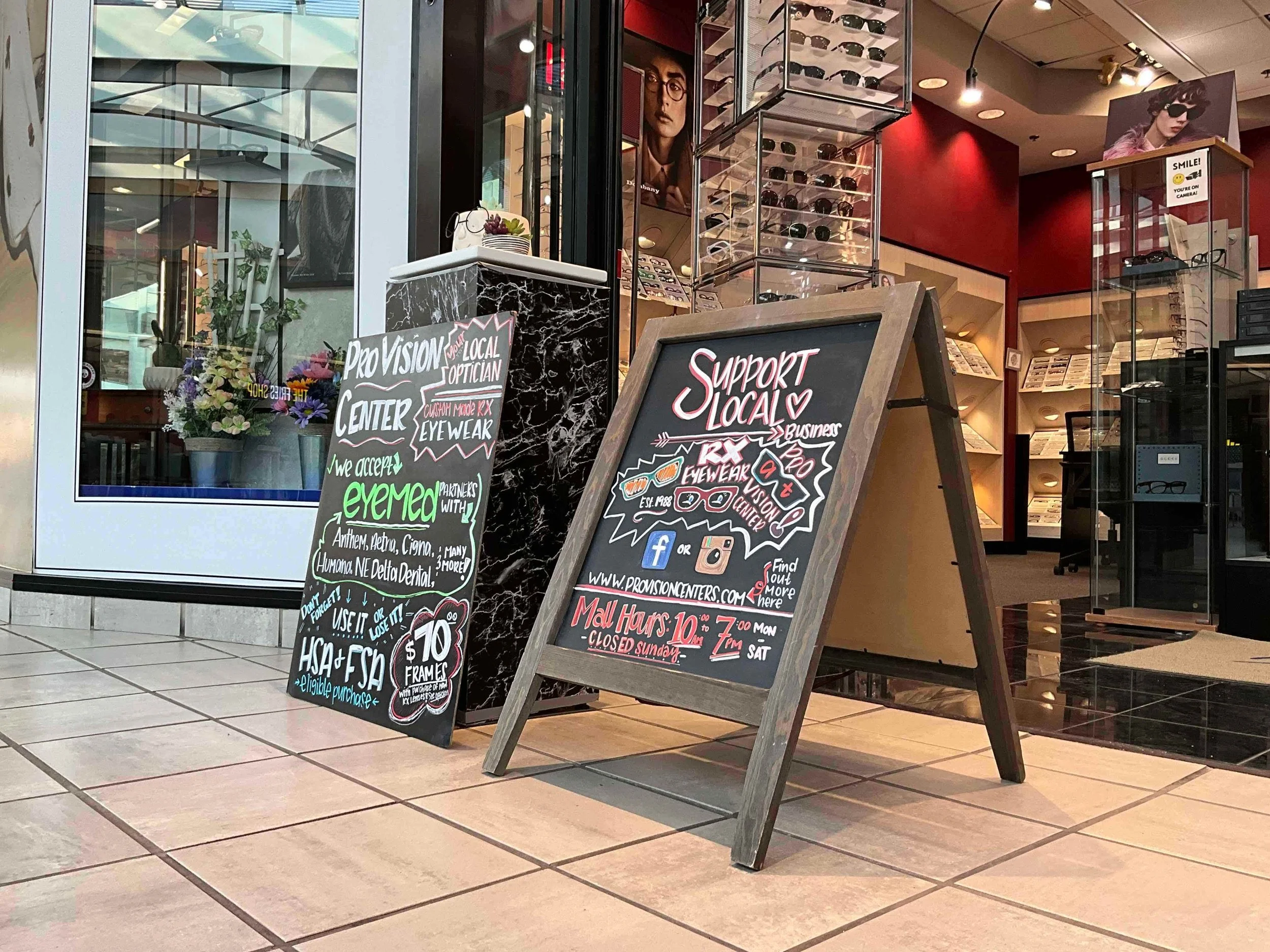 Two chalkboard signs outside an eyewear store, one with information about local optician services and the other promoting supporting local business, glasses eyewear, and store hours. The store has a display of sunglasses inside and large poster images of women wearing glasses.