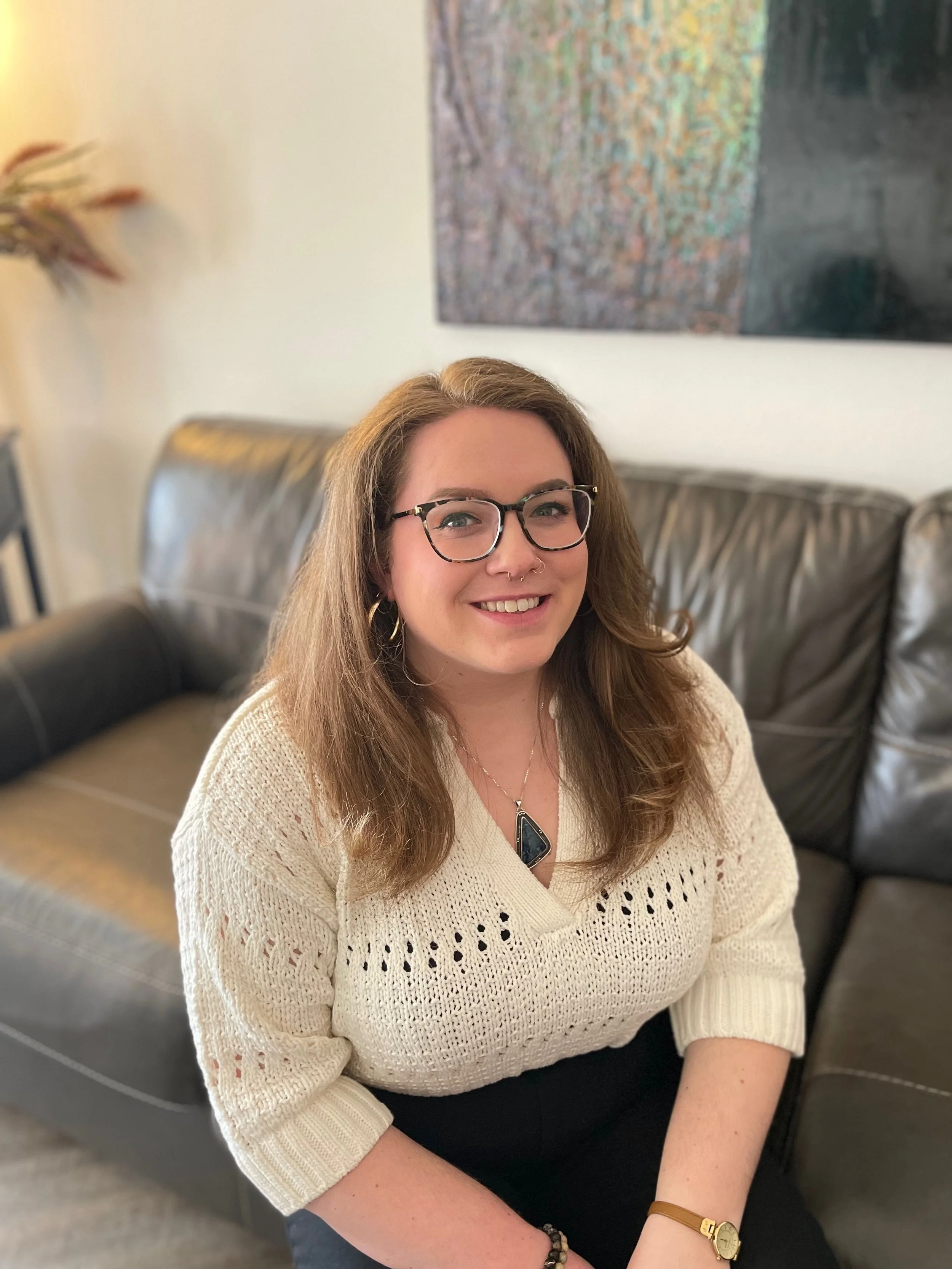 Smiling woman with glasses and curly hair standing in front of a sign that reads "The Cozy Couch Counseling & Consultation" with a drawing of a blue couch, a yellow blanket, and a plant on a small table.