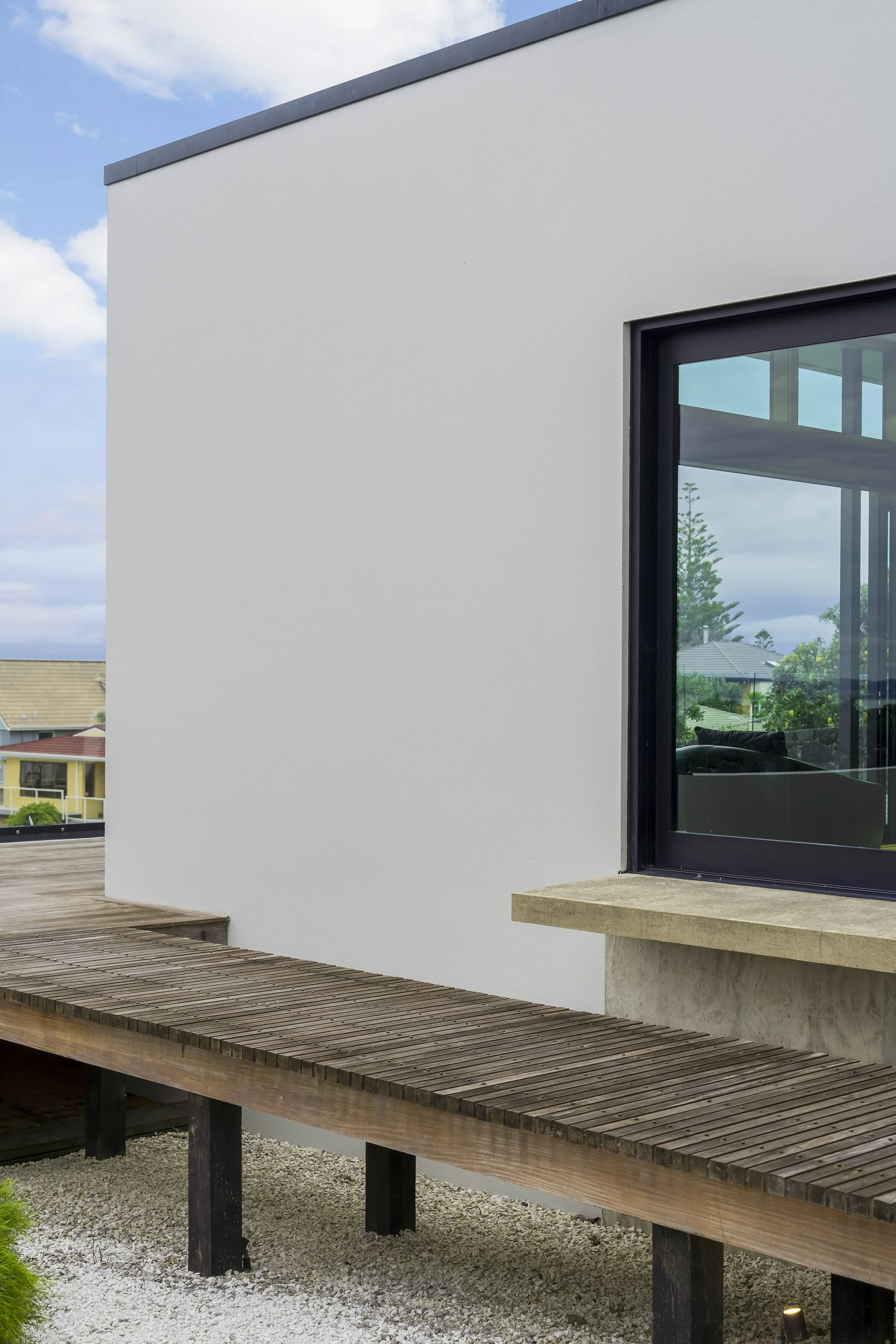 Modern house exterior with white walls and a large window, featuring a wooden deck walkway and a view of suburban rooftops and greenery.