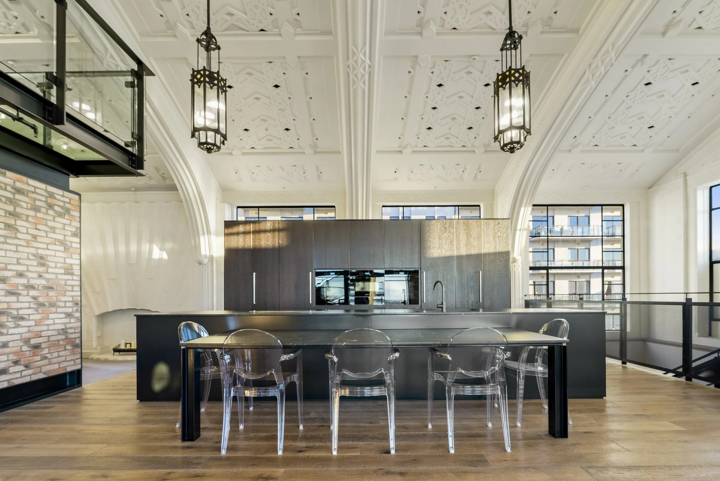 Modern kitchen interior with high ceilings featuring ornate plasterwork, large windows, a black island, transparent chairs, and hanging lantern-style lights.