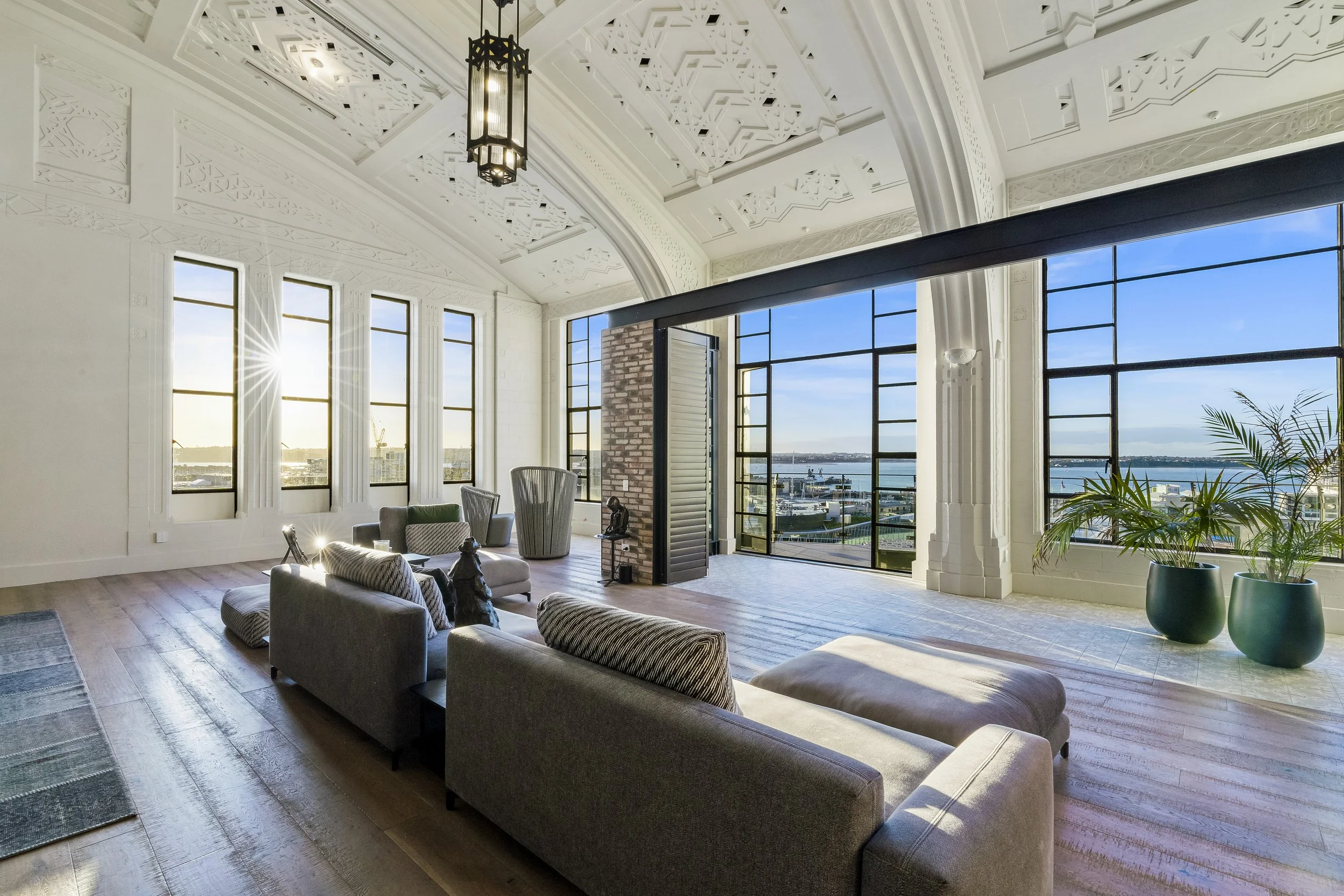 Spacious modern living room with high ceiling, large windows, and ocean view; featuring gray sofas, decorative pillows, potted plants, and intricate ceiling design.