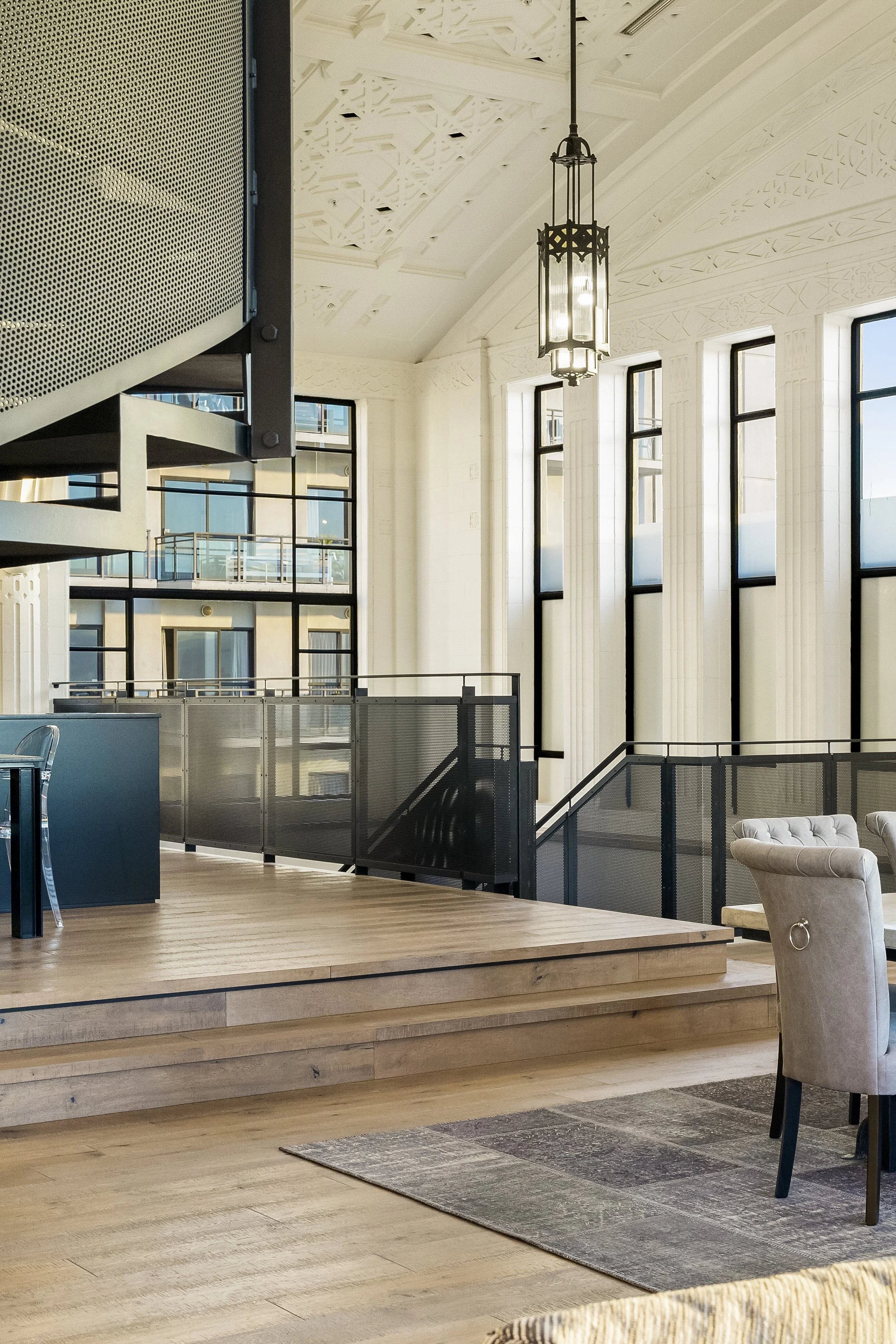 Modern interior with high ceilings, large windows, and elegant lighting; features a staircase, wooden flooring, and stylish chairs.