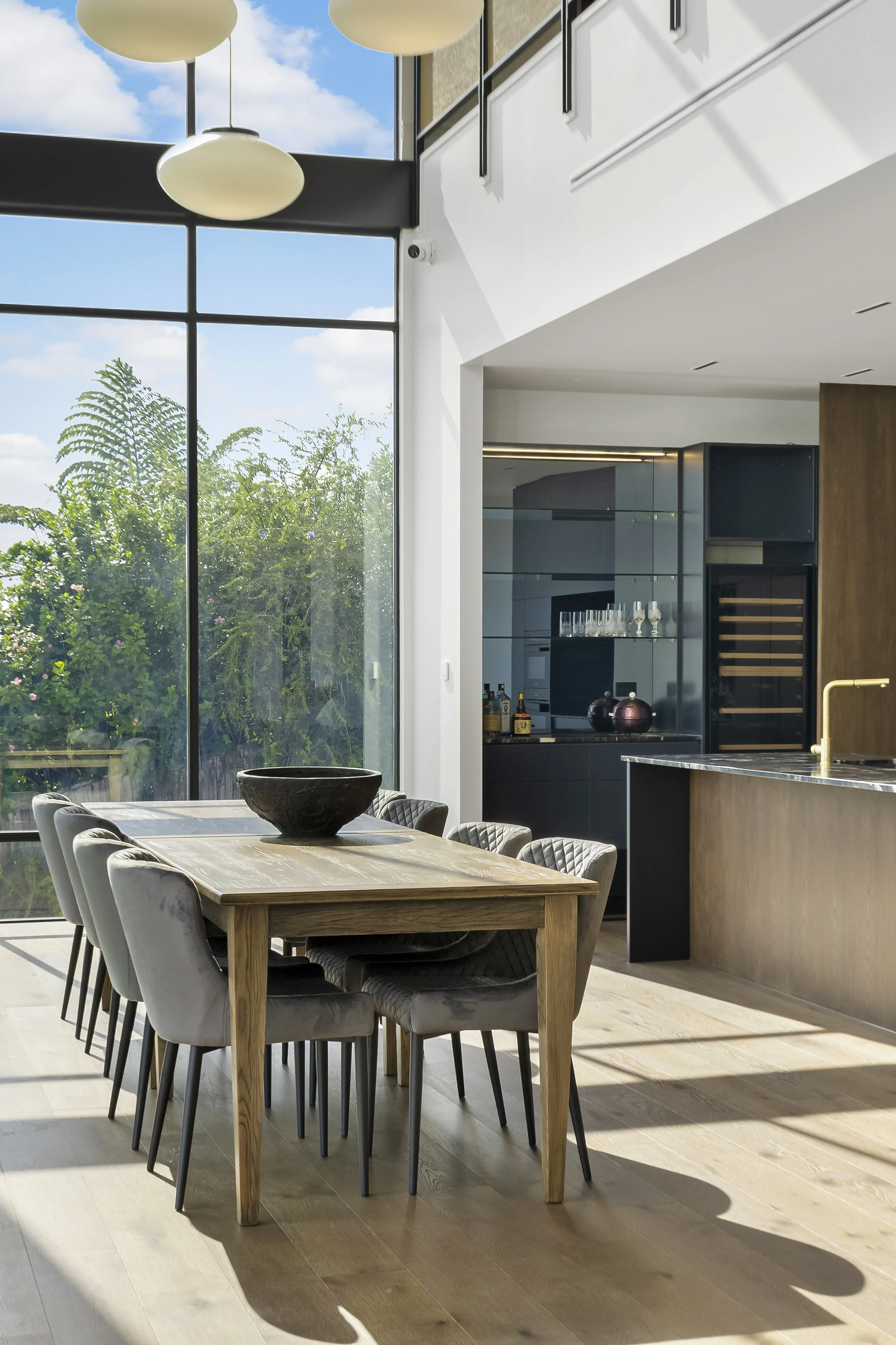 Modern dining room with a wooden table and gray chairs, large windows with greenery view, pendant lights, and a kitchen area.