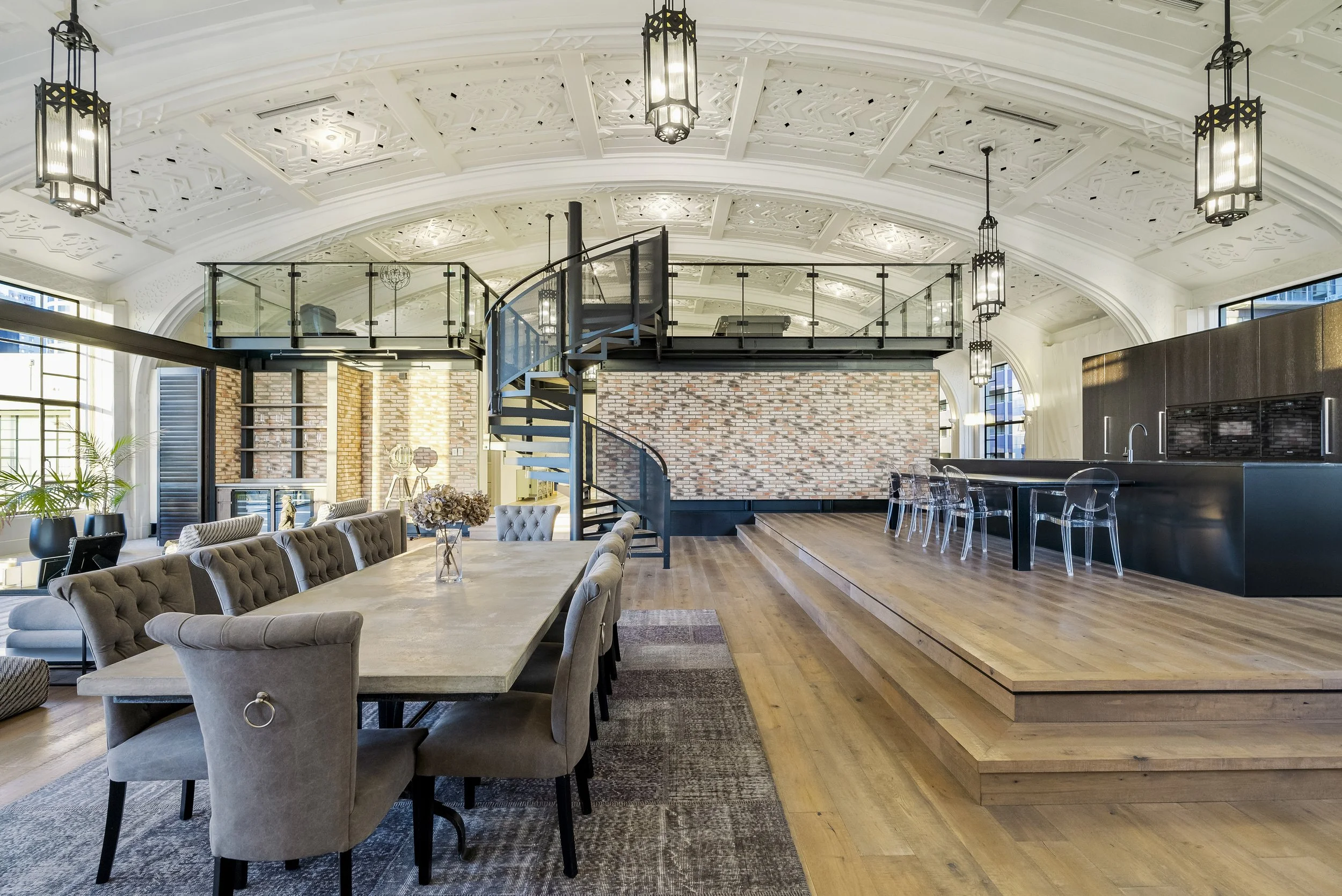 Spacious modern loft interior with high ceilings, featuring a large dining table with tufted chairs, a spiral staircase leading to an upper mezzanine, contemporary lighting fixtures, brick accent walls, and a sleek open kitchen with transparent bar s