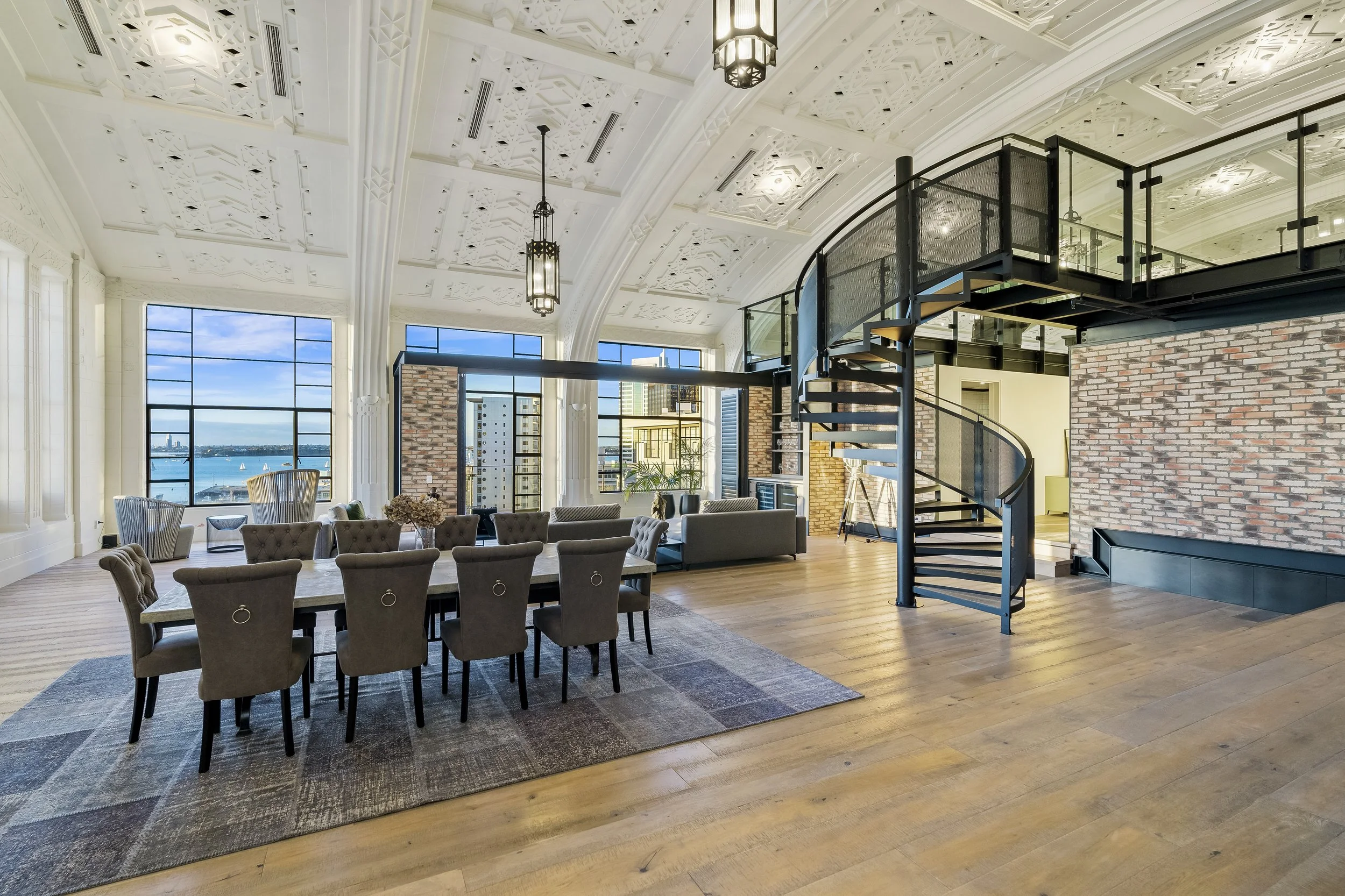 Spacious loft with a modern design, featuring a dining table with upholstered chairs, large windows with views of a city and water, spiral staircase leading to an upper level, decorative ceiling, and brick walls.