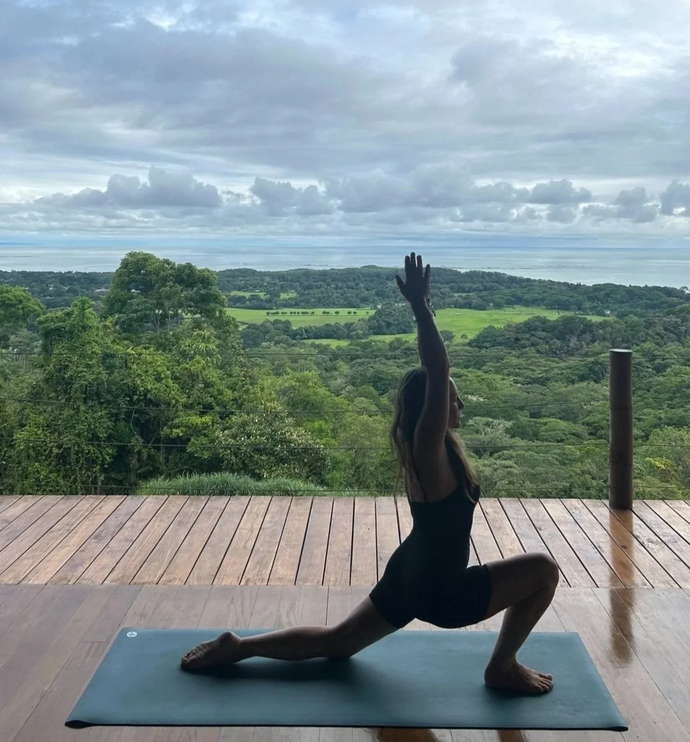Woman practicing yoga outdoors on a mat, performing a lunge with arms raised, overlooking a lush, green forested landscape with the ocean and a cloudy sky in the background in Uvita, Costa Rica.